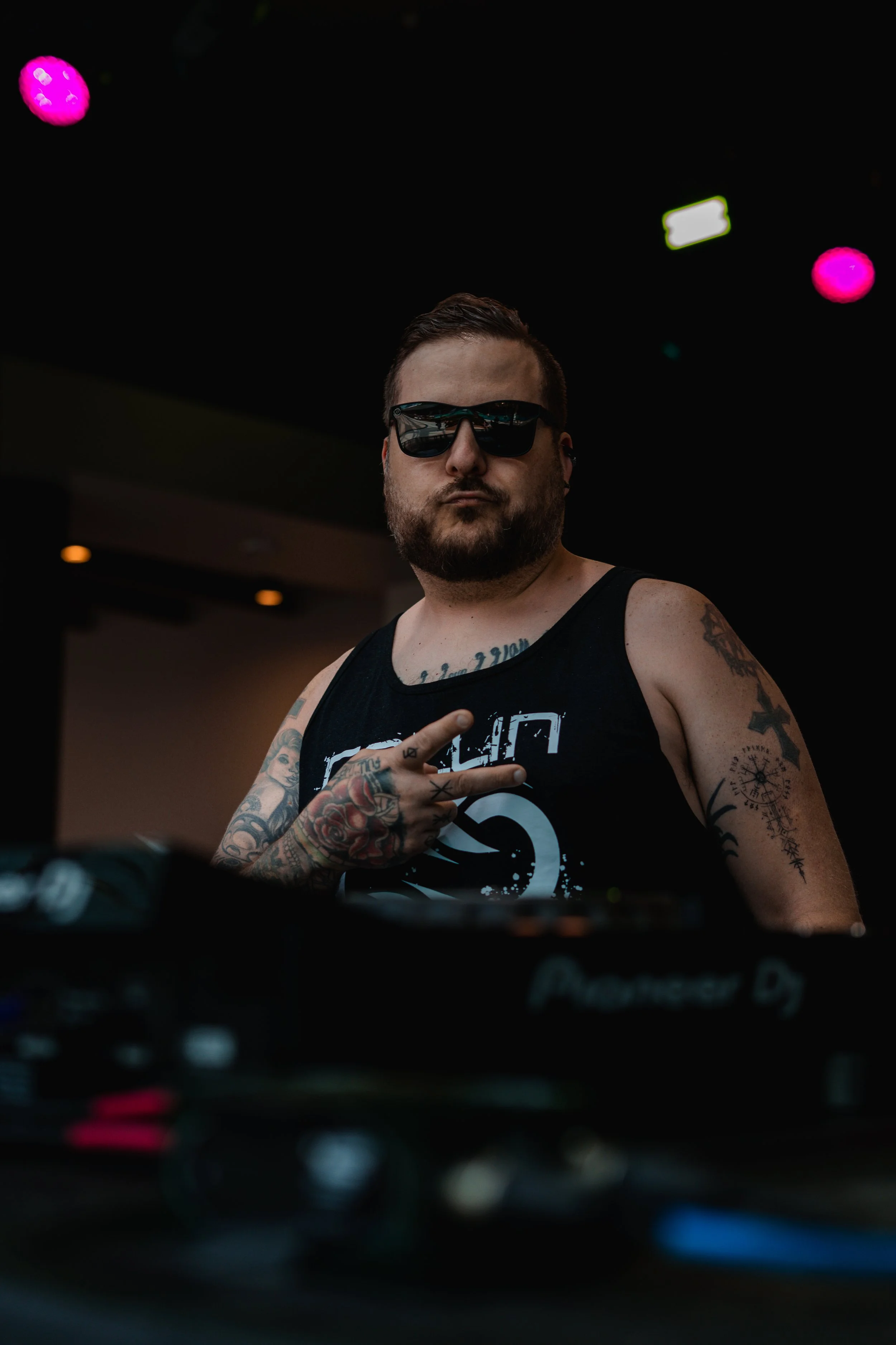 A DJ with tattoos, wearing sunglasses and a black tank top, standing collin chaos console in a dark club environment. artist prominent dj grand rapids ready for bookings and big festivals, artist, founder remedy collective djs sound production