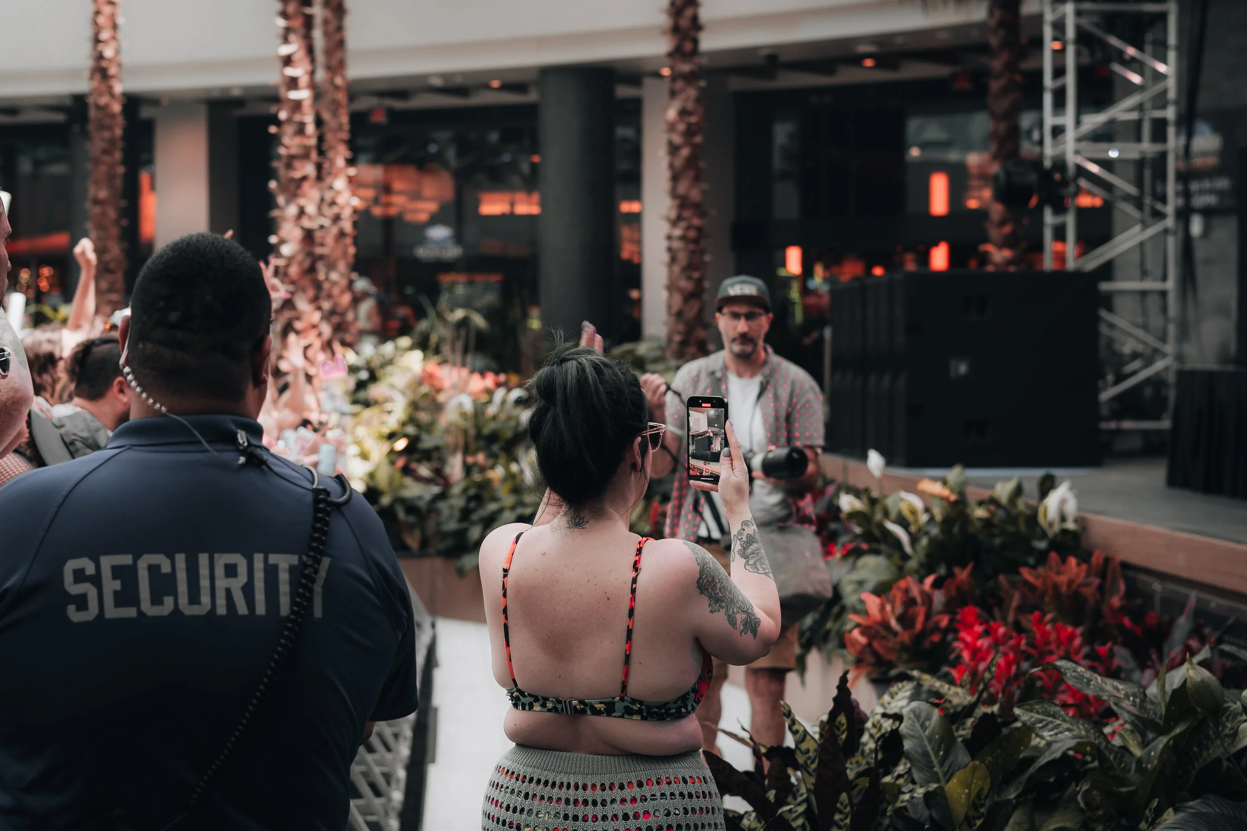 A woman with tattoos on her arm is taking a selfie with a man in a patterned shirt and cap at an indoor event with floral decorations, security personnel, and a stage in the background.