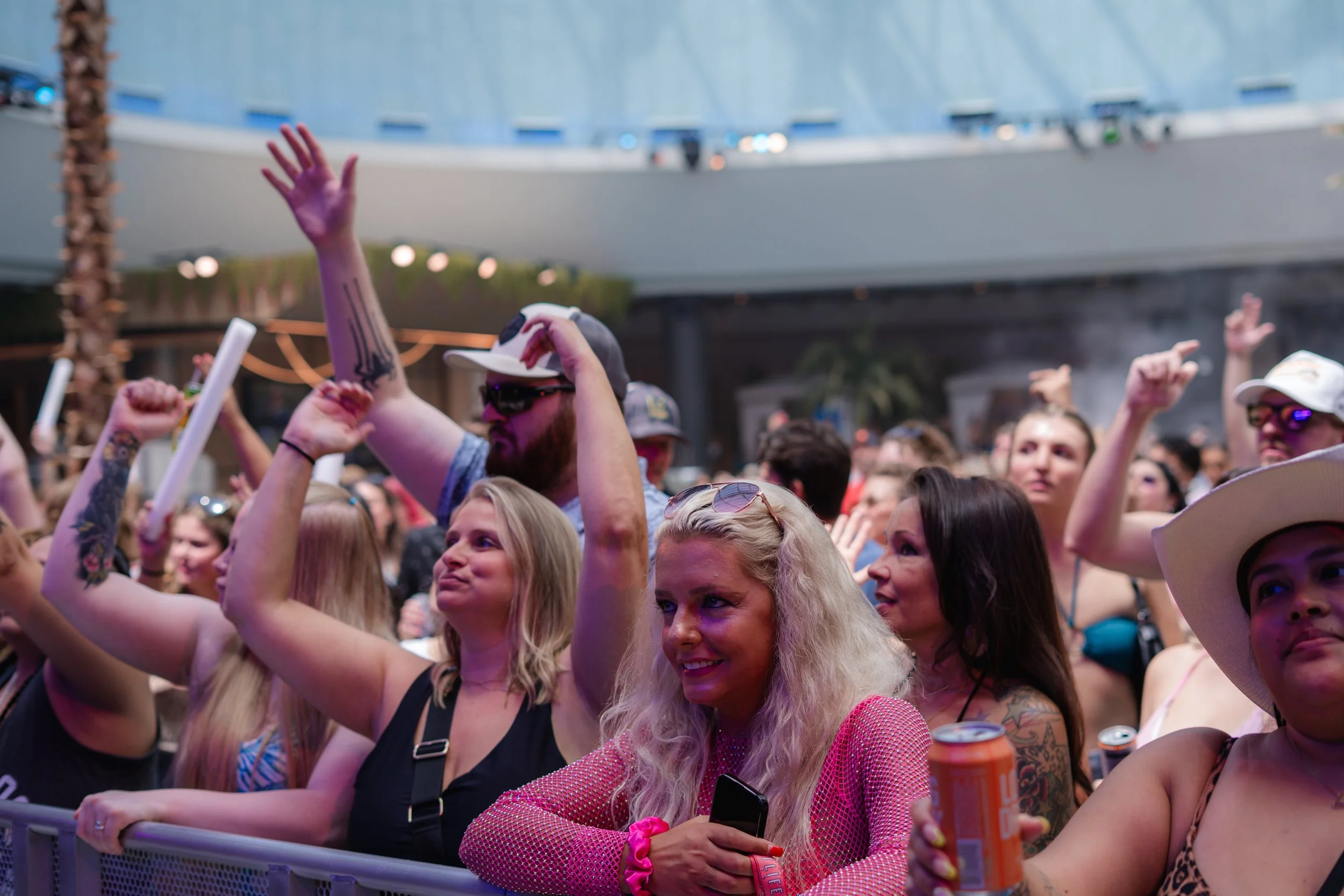 People at an outdoor concert or festival enjoying the event, some raising their hands and holding drinks, with a stage or performance area in the background.