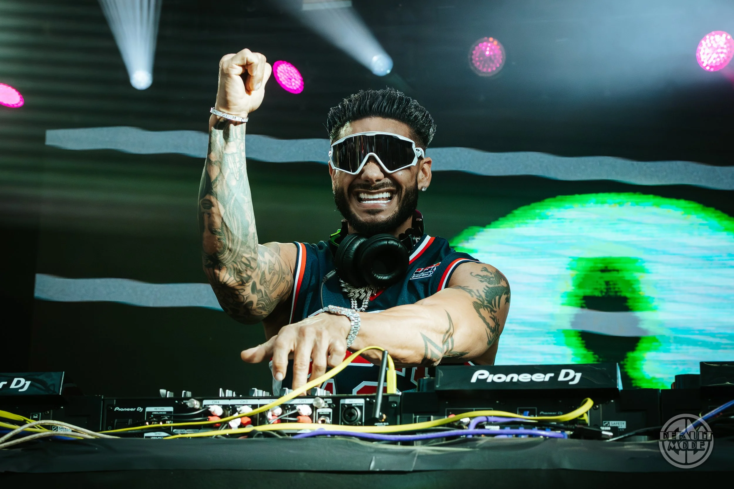A DJ with sunglasses and headphones around his neck, smiling and raising his arm, performing at a nightclub or concert with colorful lights in the background.