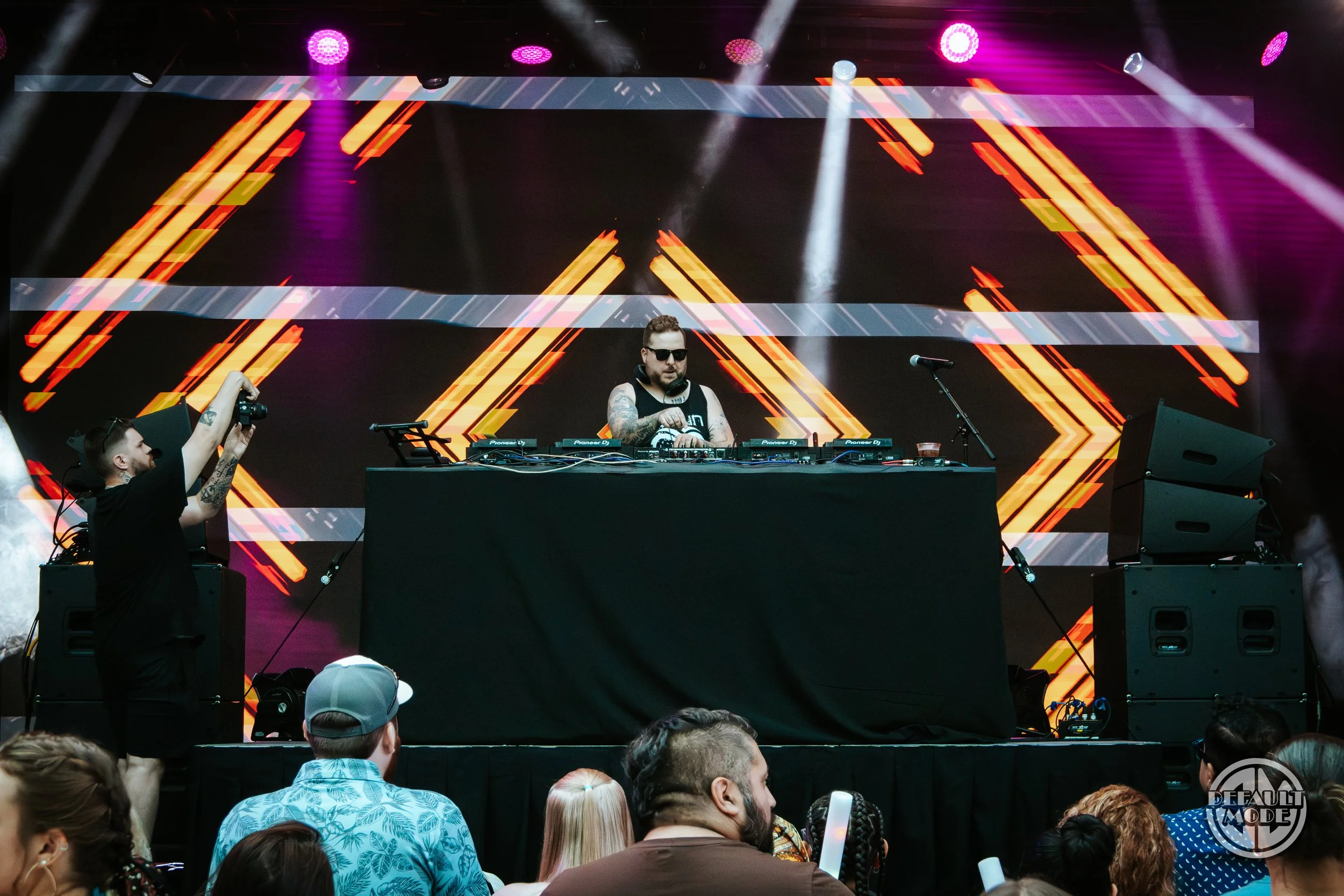 DJ performing on stage with colorful digital visuals behind him, audience watching, and a photographer taking pictures.