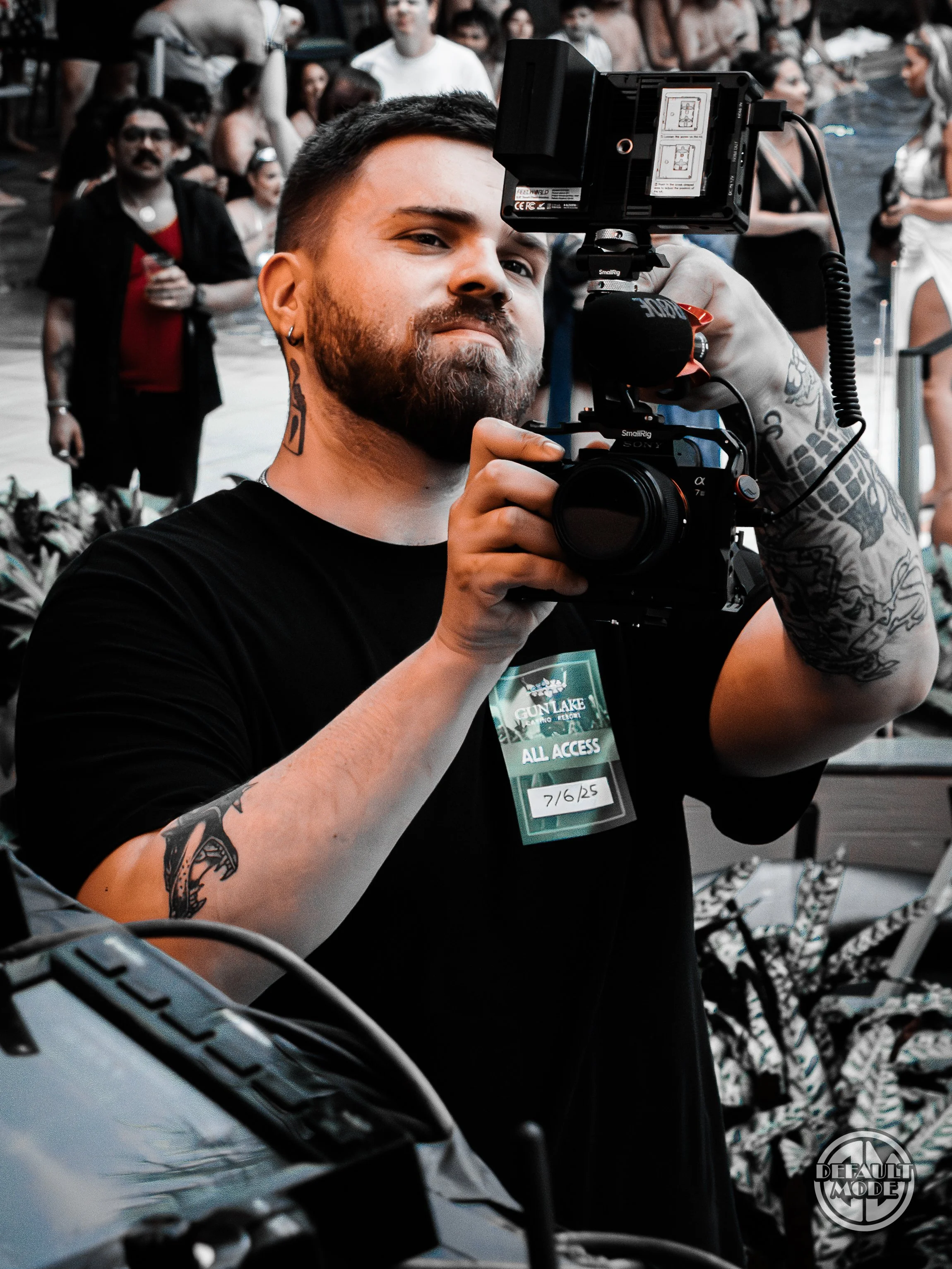 A man with a beard, tattoos, and a black t-shirt is taking photos with a professional camera at an outdoor event with a crowd of people in the background.