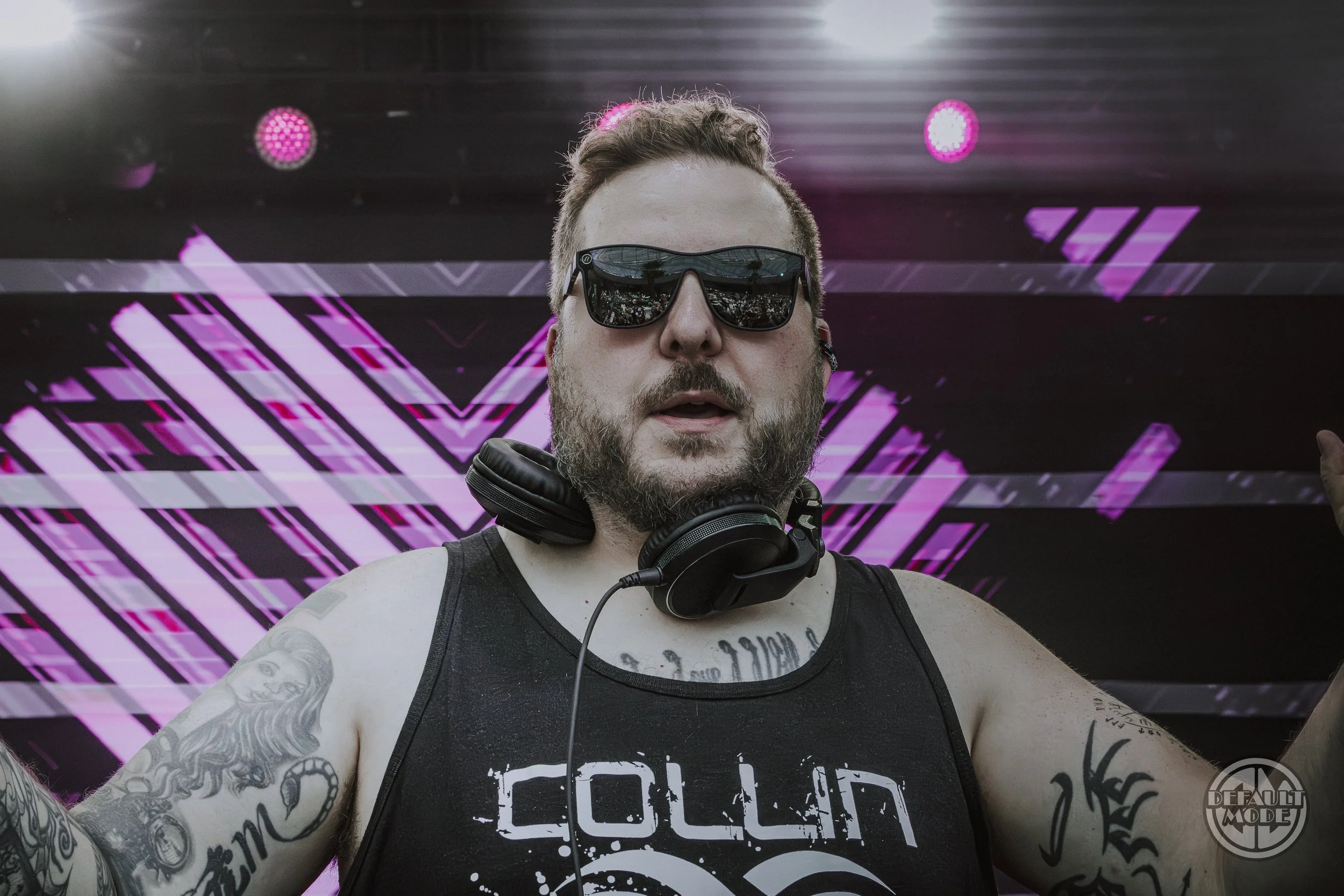 A bearded man wearing sunglasses, headphones around his neck, a tank top, and tattoos, standing in front of a colorful digital backdrop at a music event.
