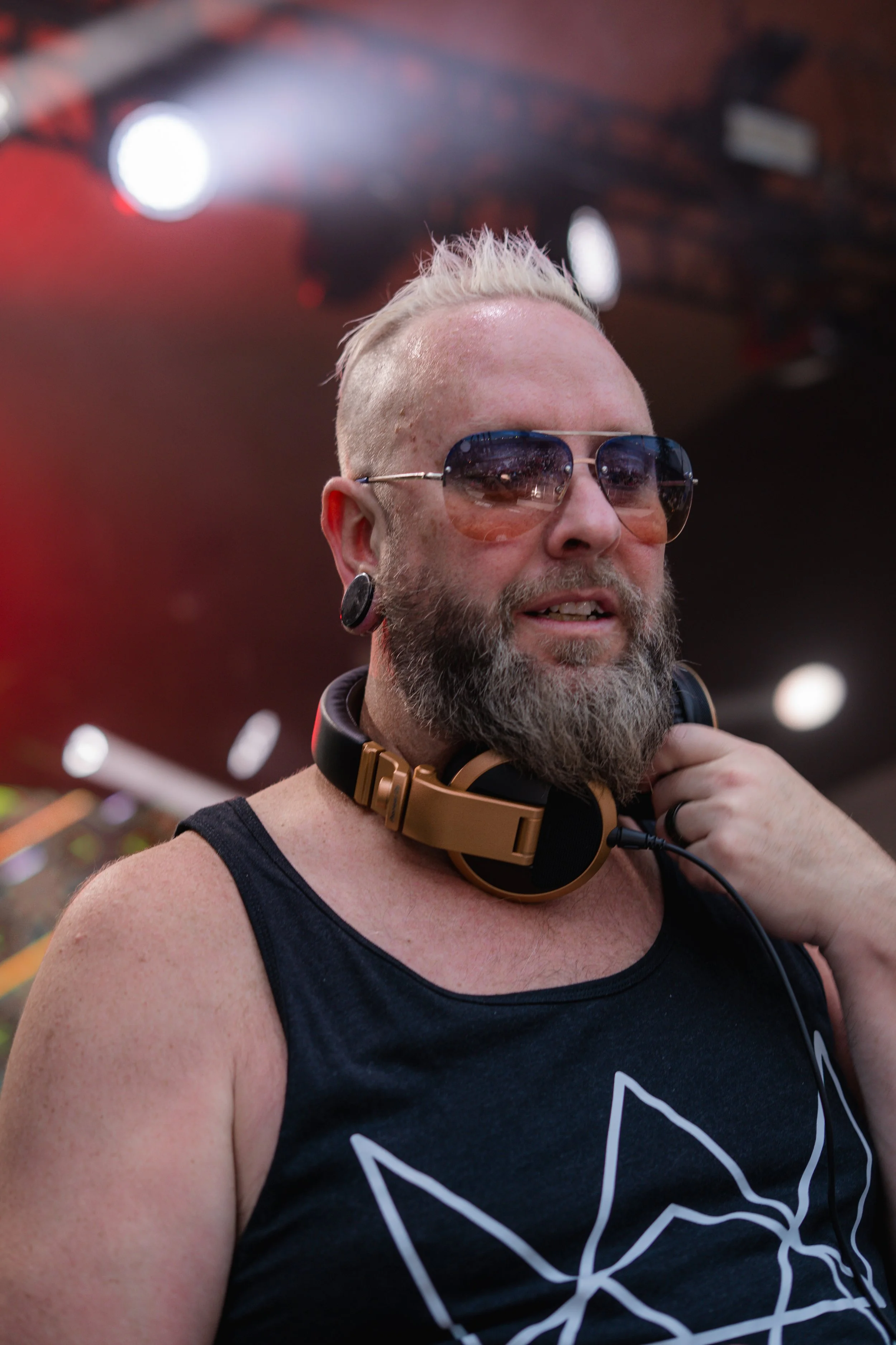 A man with a beard and sunglasses, wearing a tank top and over-ear headphones, at a music event. mattb artist prominent dj grand rapids ready for bookings and big festivals, artist, founder remedy collective djs sound production lighting mattb tours