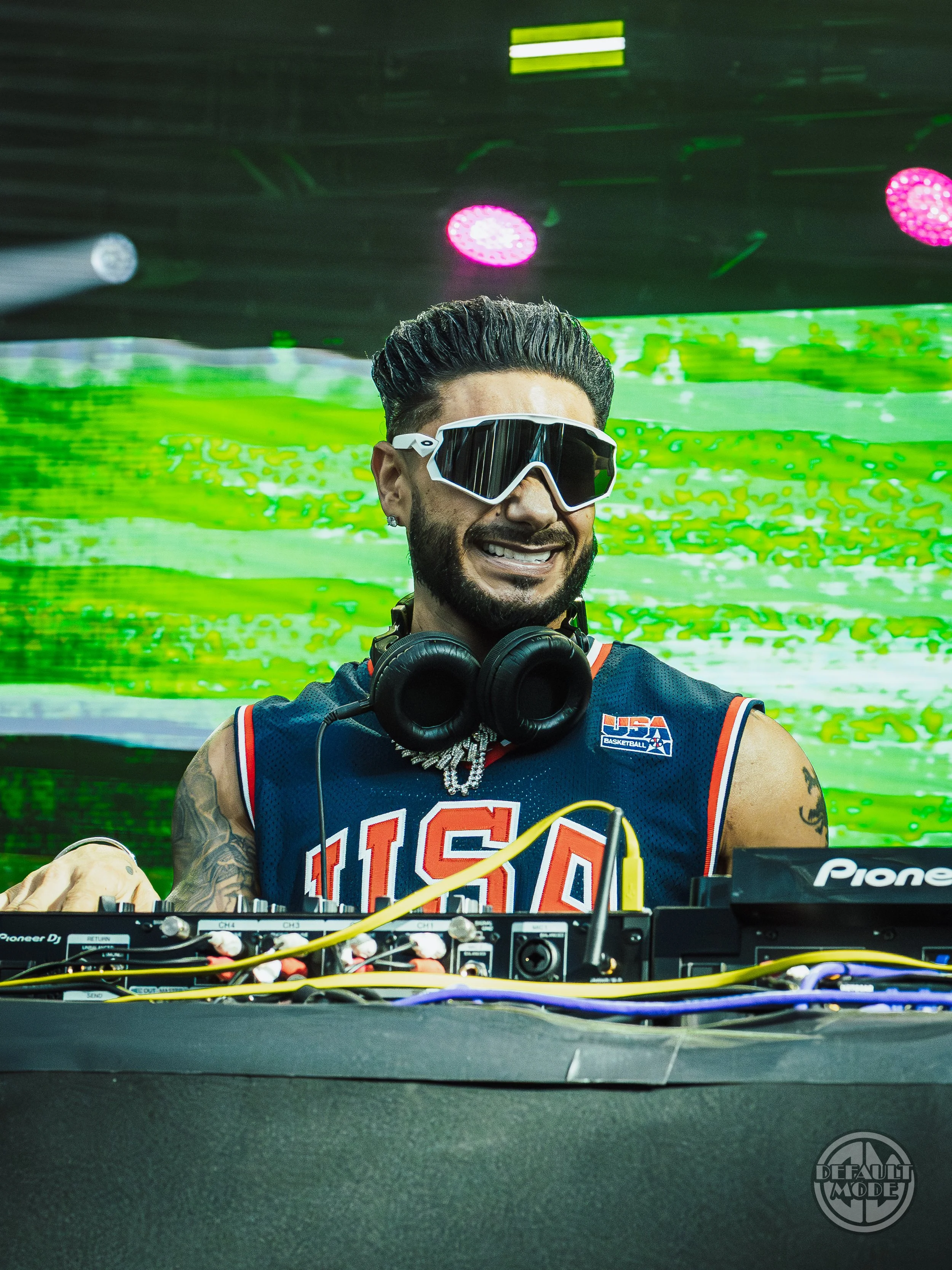 A male DJ wearing sunglasses, a USA basketball jersey, and headphones around his neck, smiling while performing at a music event with colorful lighting and a green digital background.