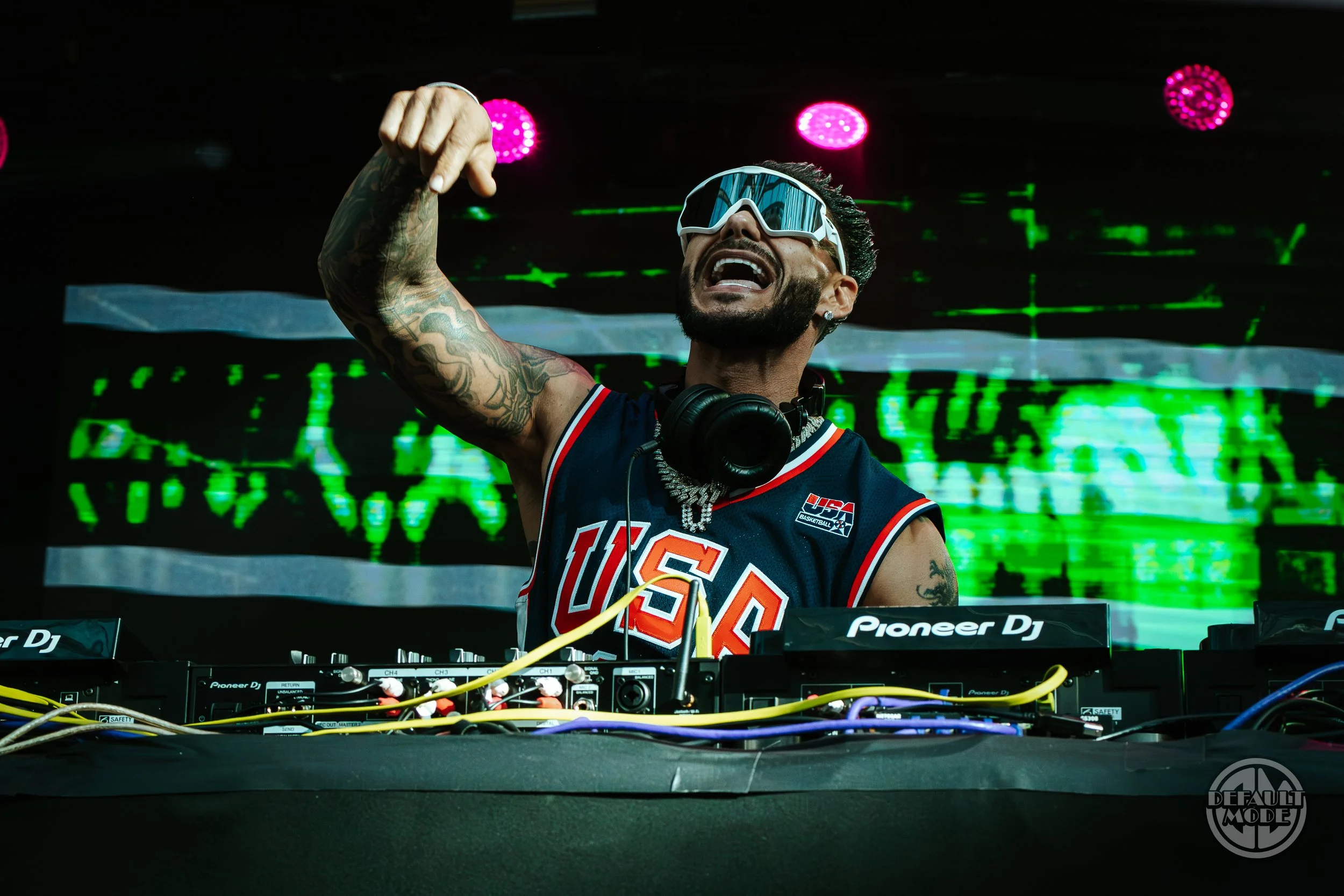 DJ in a USA basketball jersey performing as a DJ at a concert, wearing sunglasses and headphones with a digital green backdrop.