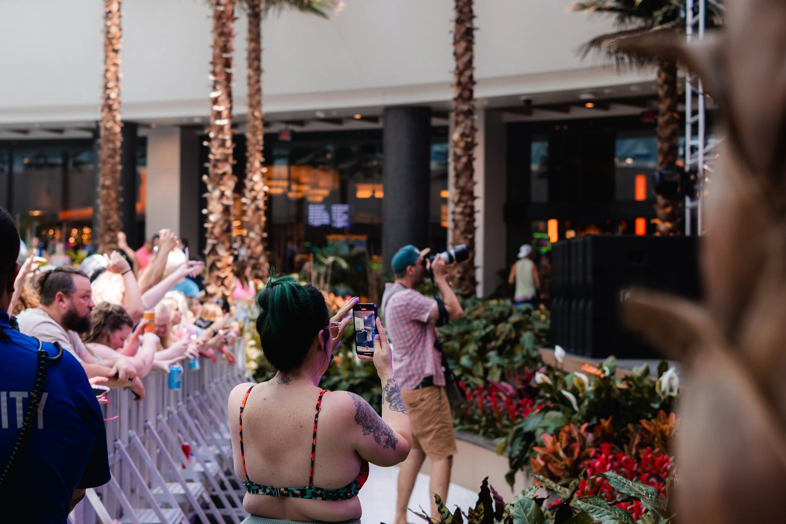 A crowd of people taking photos and videos at an outdoor event, with some standing behind a barrier and others holding up devices, in front of a stage with palm trees in the background.