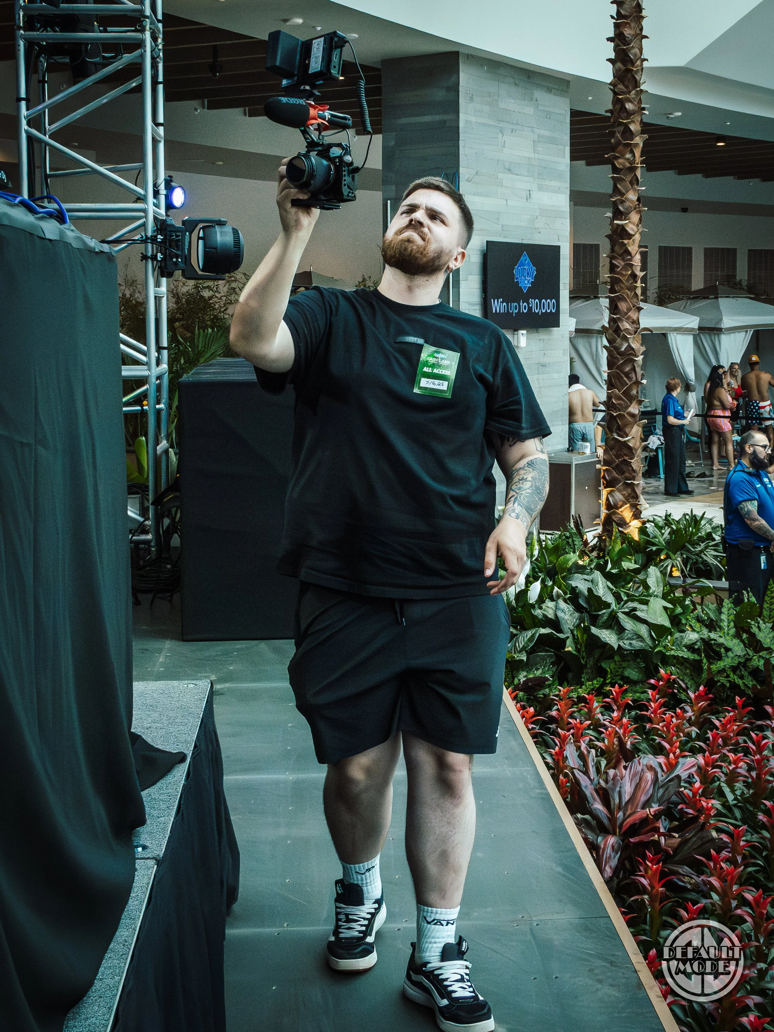 A man with a beard and tattoos on his left arm is holding a video camera, standing on a stage with a dark curtain to his left and plants to his right. He has a serious expression, is wearing a black T-shirt, black shorts, white socks, and black sneak