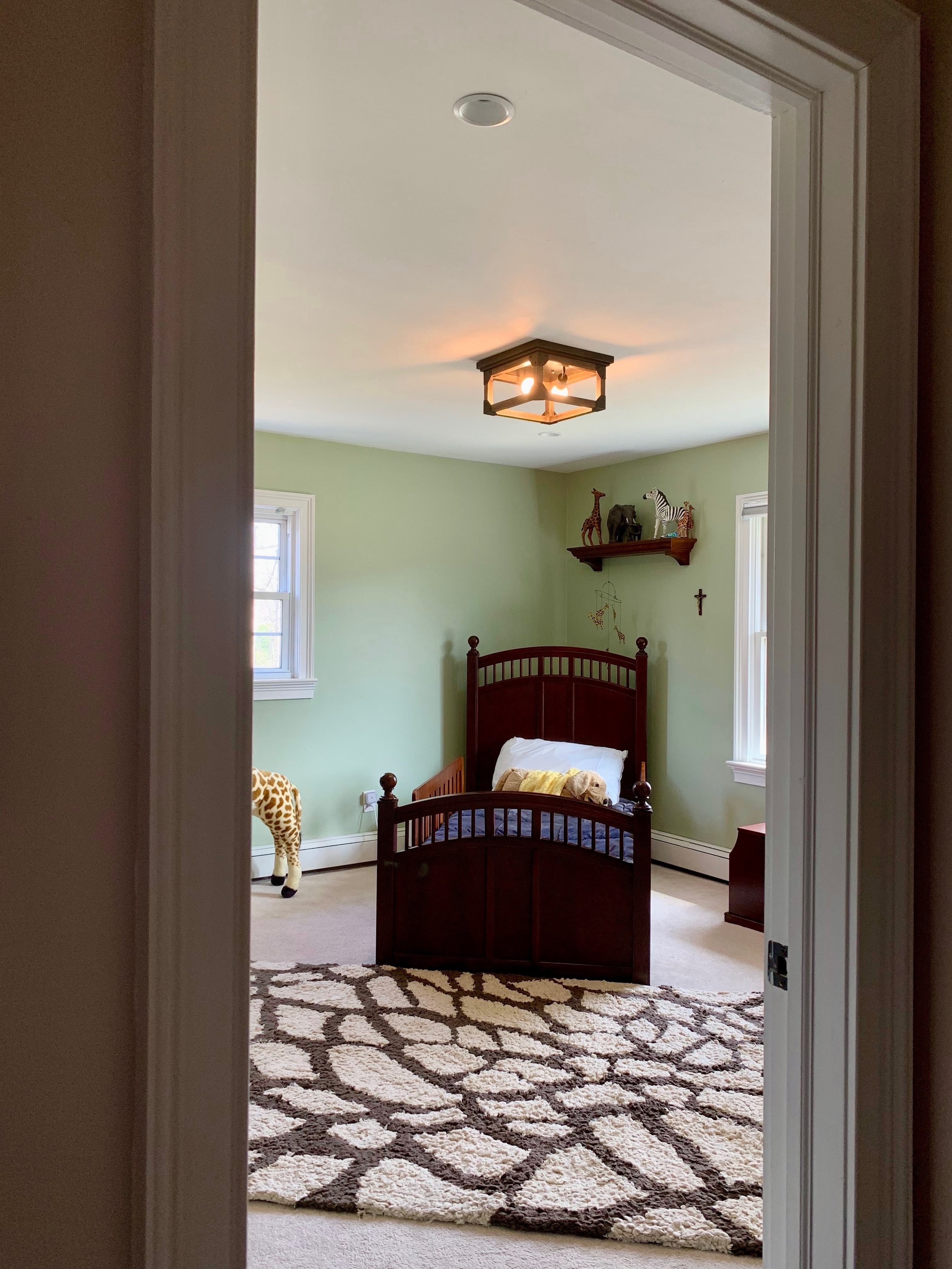 A child's bedroom with green walls, a wooden bed with a pillow and stuffed animal, a shelf with animal figurines, a giraffe stuffed toy on the floor, a patterned rug, and two windows.