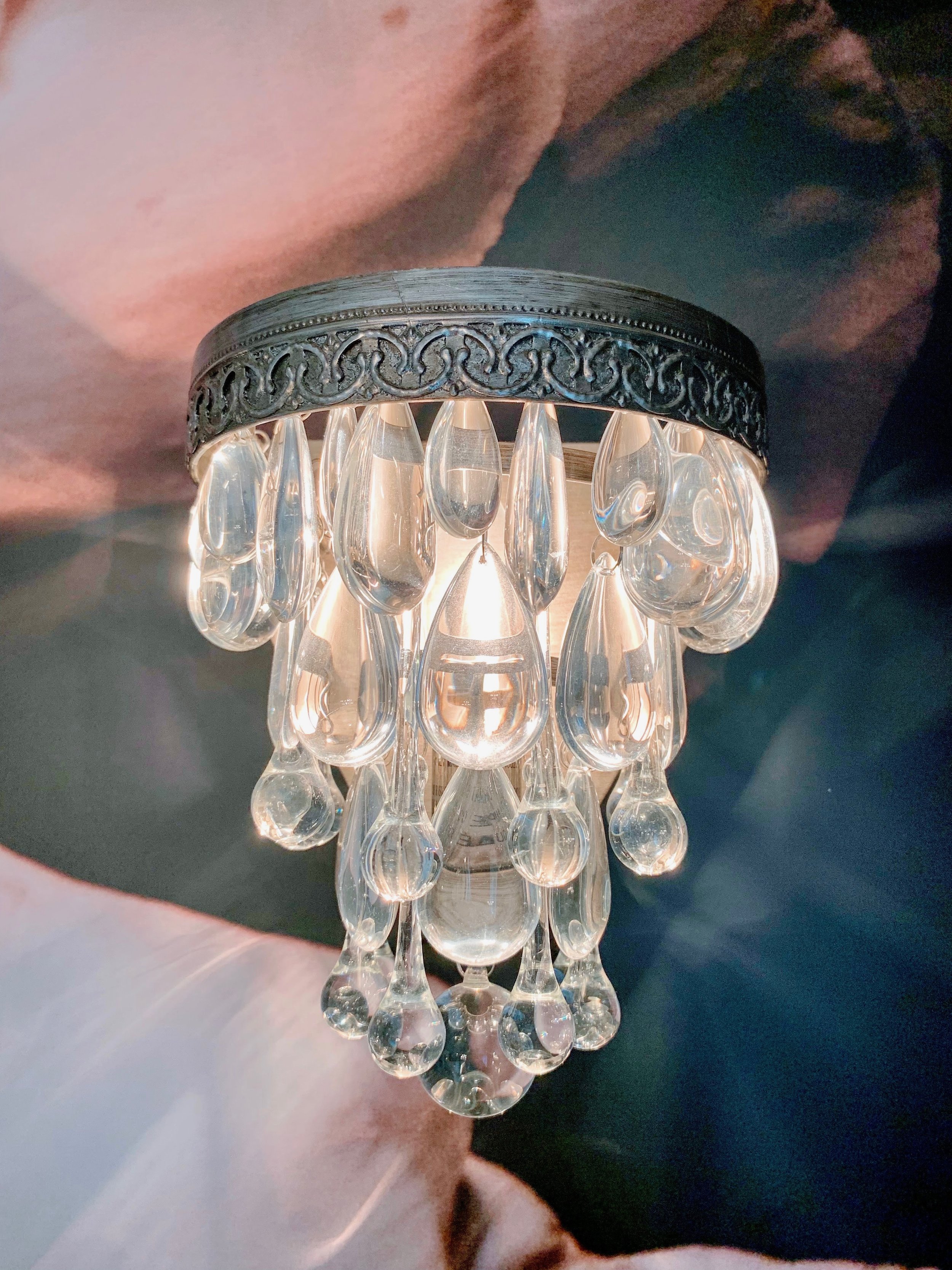 Close-up of a small chandelier with hanging glass droplets, mounted on a wall with a partial blurred face portrait in the background.