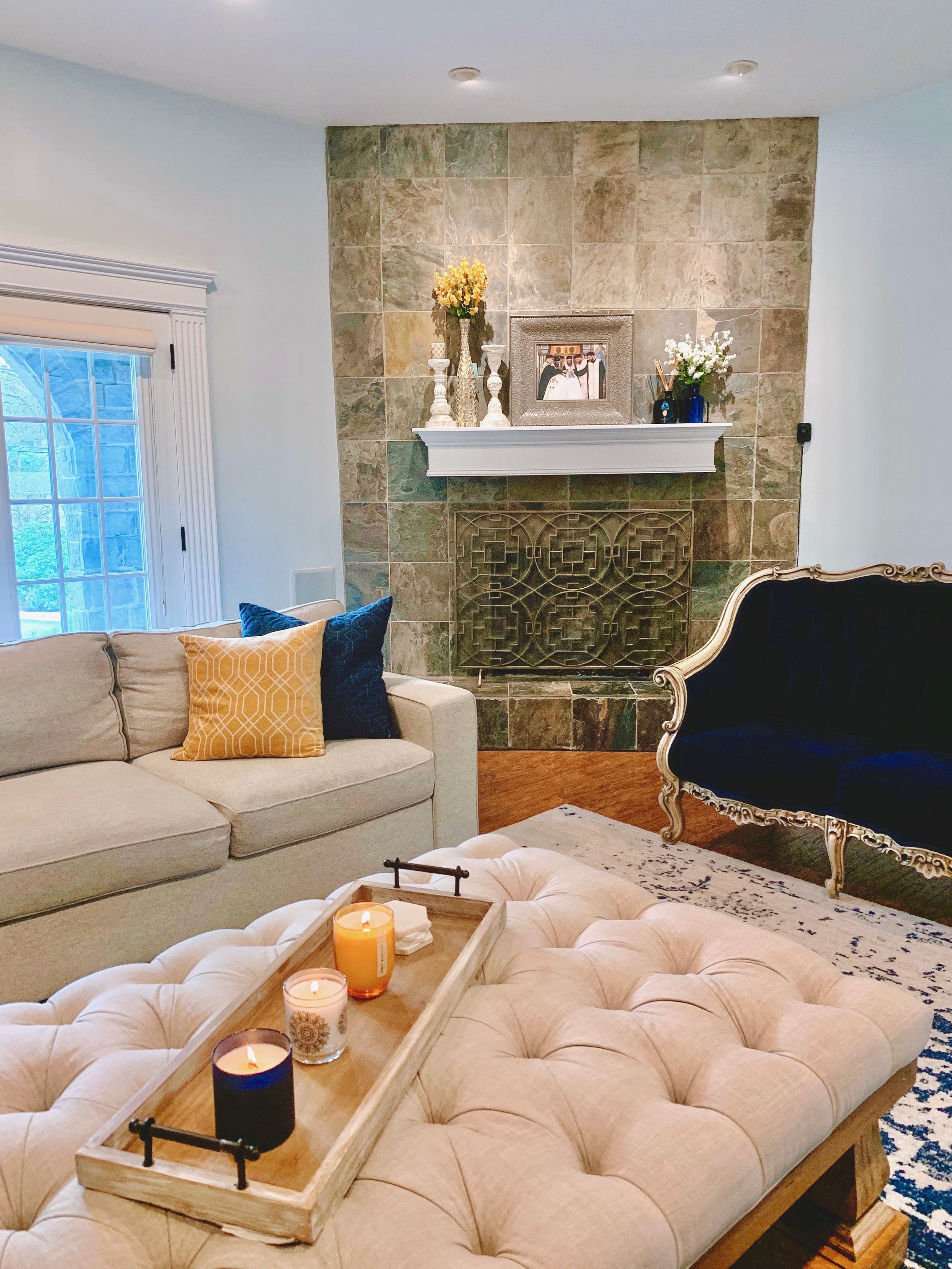 A cozy living room featuring a stone fireplace with decorative items on the mantel, a beige tufted ottoman with candles in a wooden tray, a light-colored sofa with throw pillows, a dark blue vintage-style armchair, and a window with white trim.