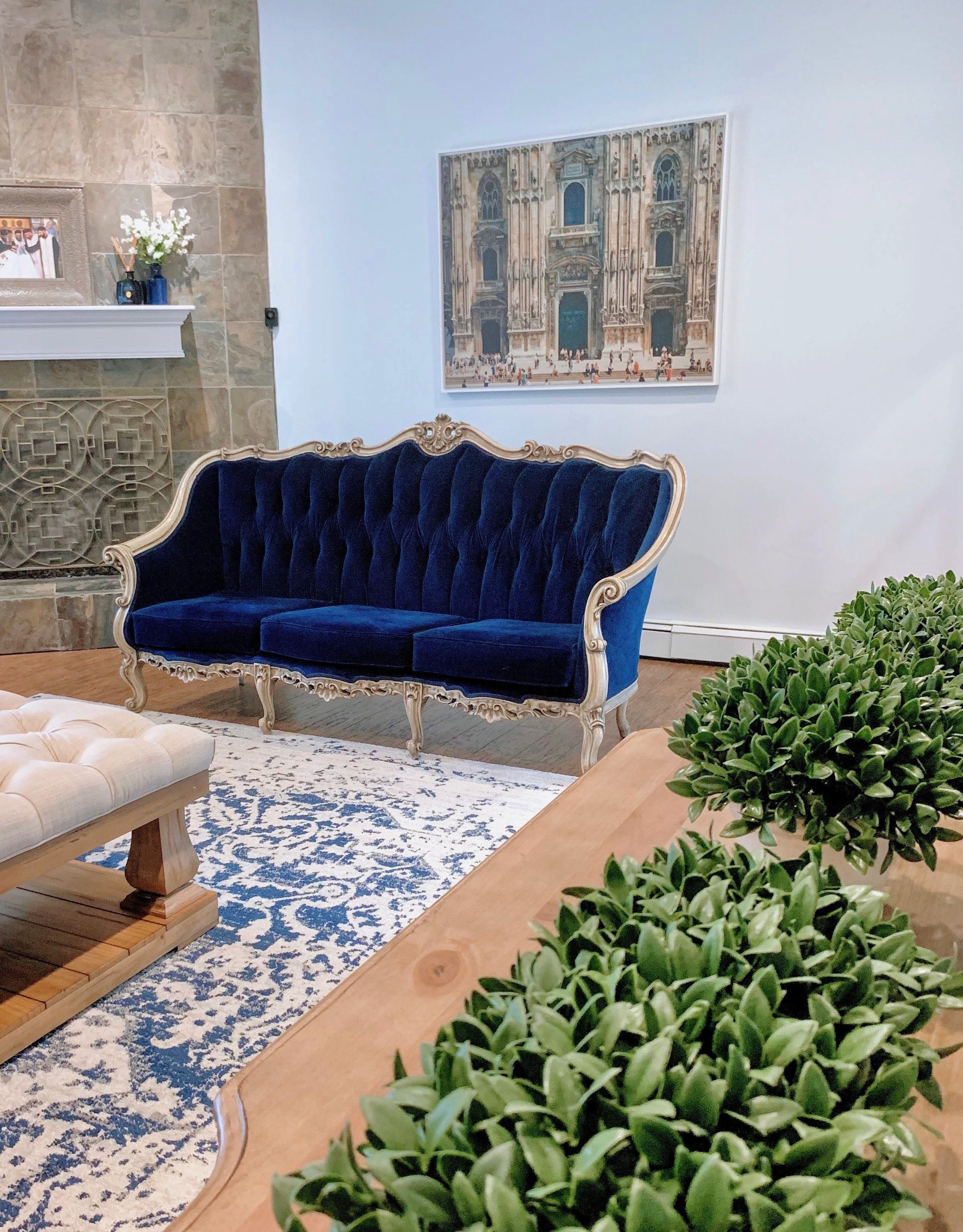 A living room with a vintage blue velvet sofa, a white and blue patterned rug, a cream-colored tufted ottoman, a wooden table with greenery, a framed picture on the wall, a fireplace with a floral arrangement on the mantel, and a large painting of a