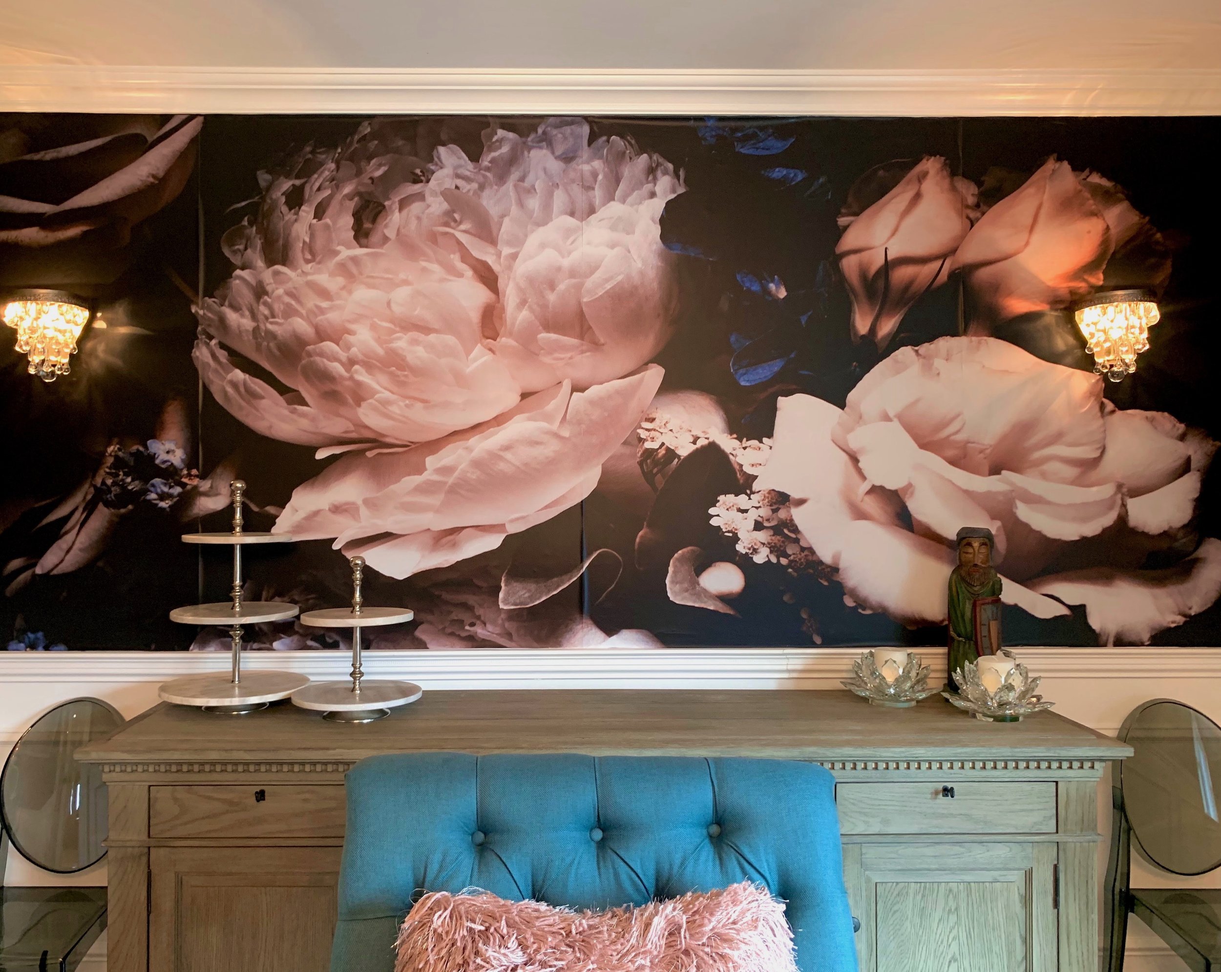 Living room with a large floral mural featuring peonies in pink and white shades on the wall behind a wooden sideboard. On the sideboard are two tiered stands, decorative glass candle holders, and a statue of a figure in a robe. A blue upholstered ch