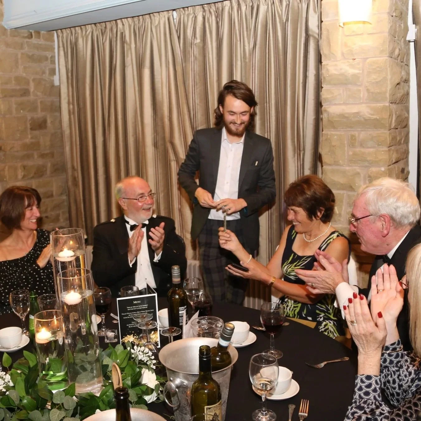 A group of older adults and one younger man at a dinner party, sitting around a table with wine, candles, and floral centerpieces, with a man standing and receiving applause.