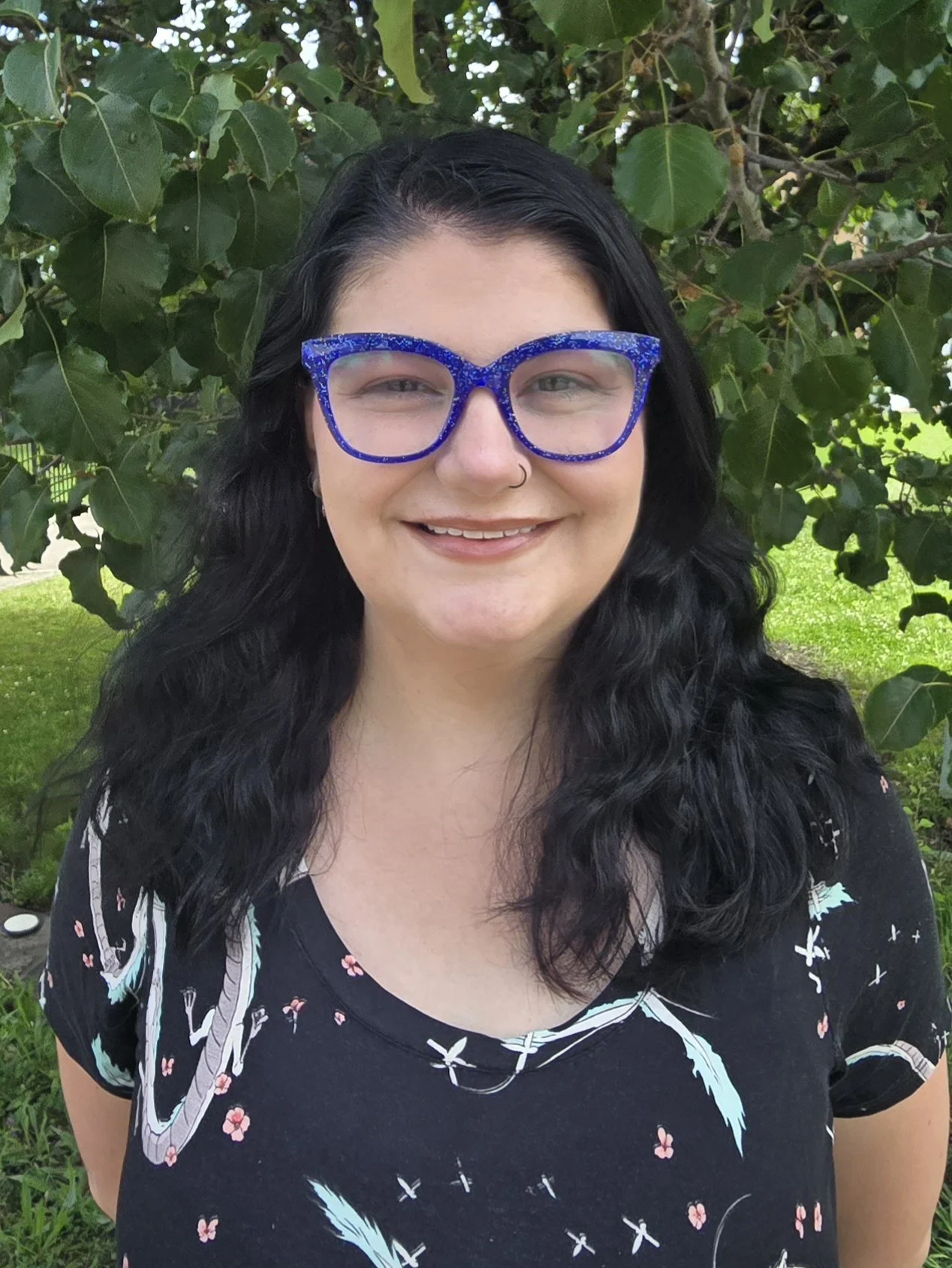 A woman with black wavy hair wearing blue glittery glasses, a black dress with white and pink floral patterns, and a nose ring, standing outdoors in front of green leafy trees.