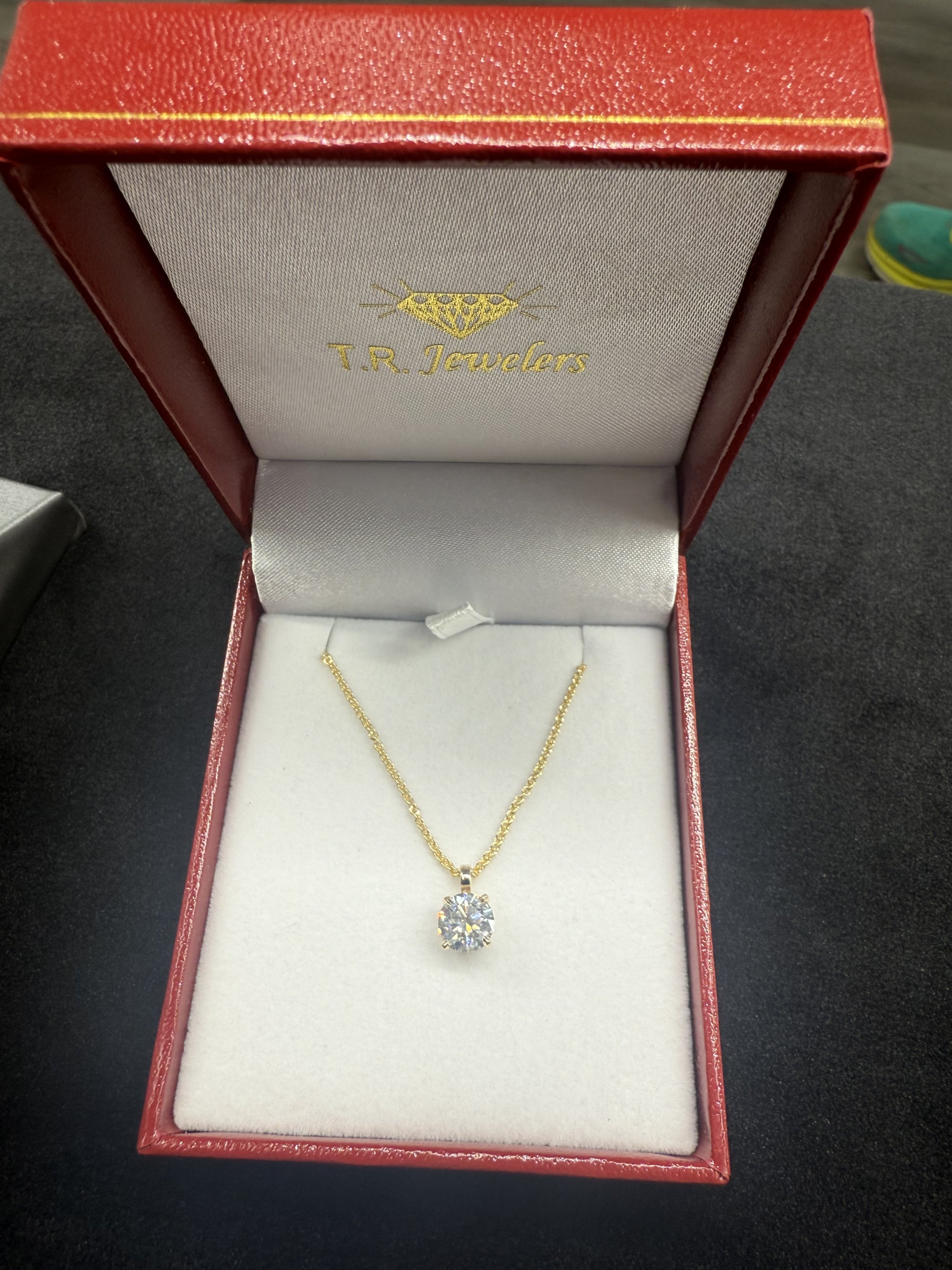 Gold necklace with a solitaire diamond pendant in a red jewelry box labeled 'T.R. Jewelers' on a black surface.