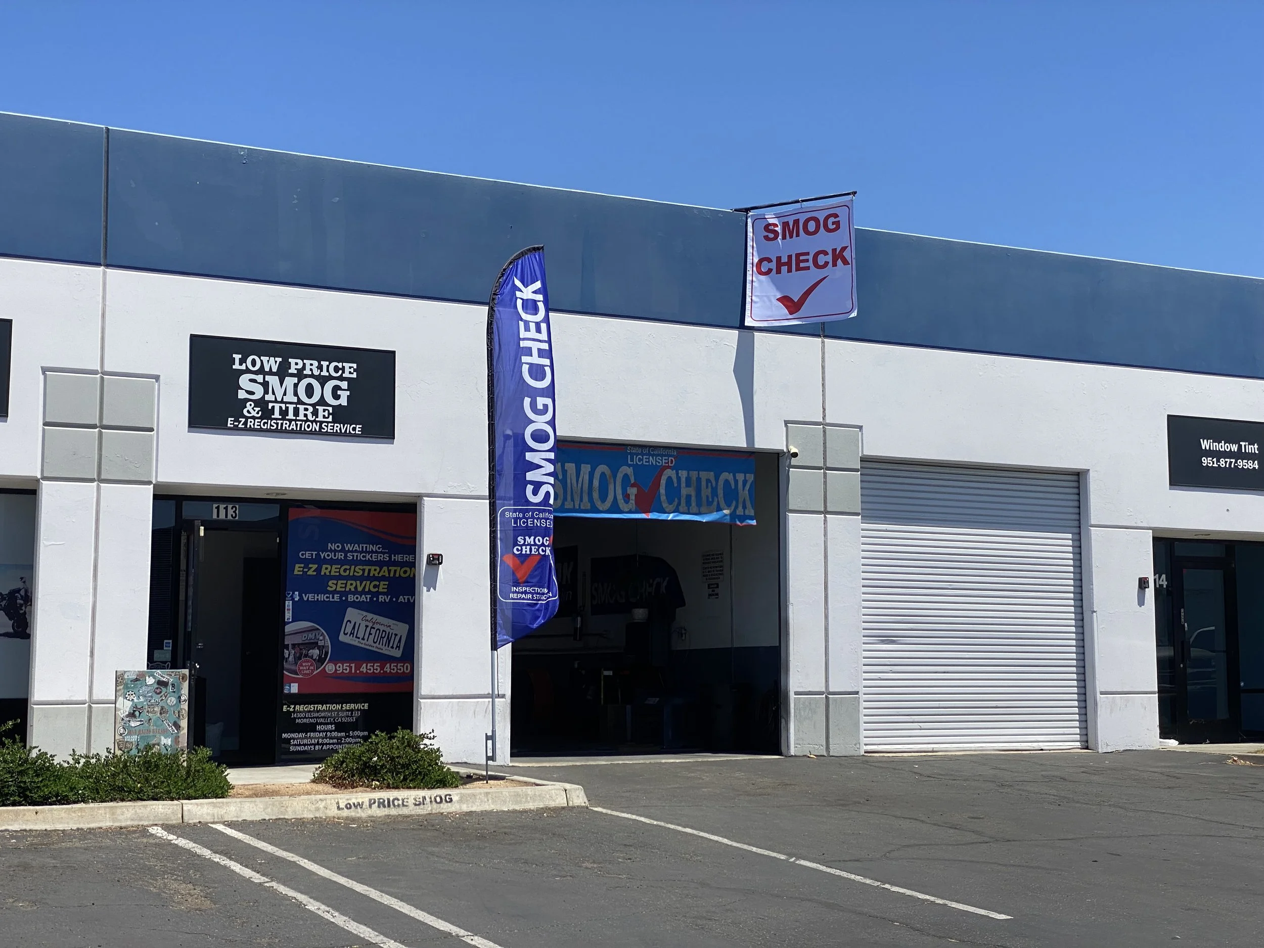 A building with signage advertising smog check, low price smog and tire services, and E-Z registration service. There are flags and banners promoting smog checks and inspections, a garage door, and a parking lot with marked parking spaces in the foreground.