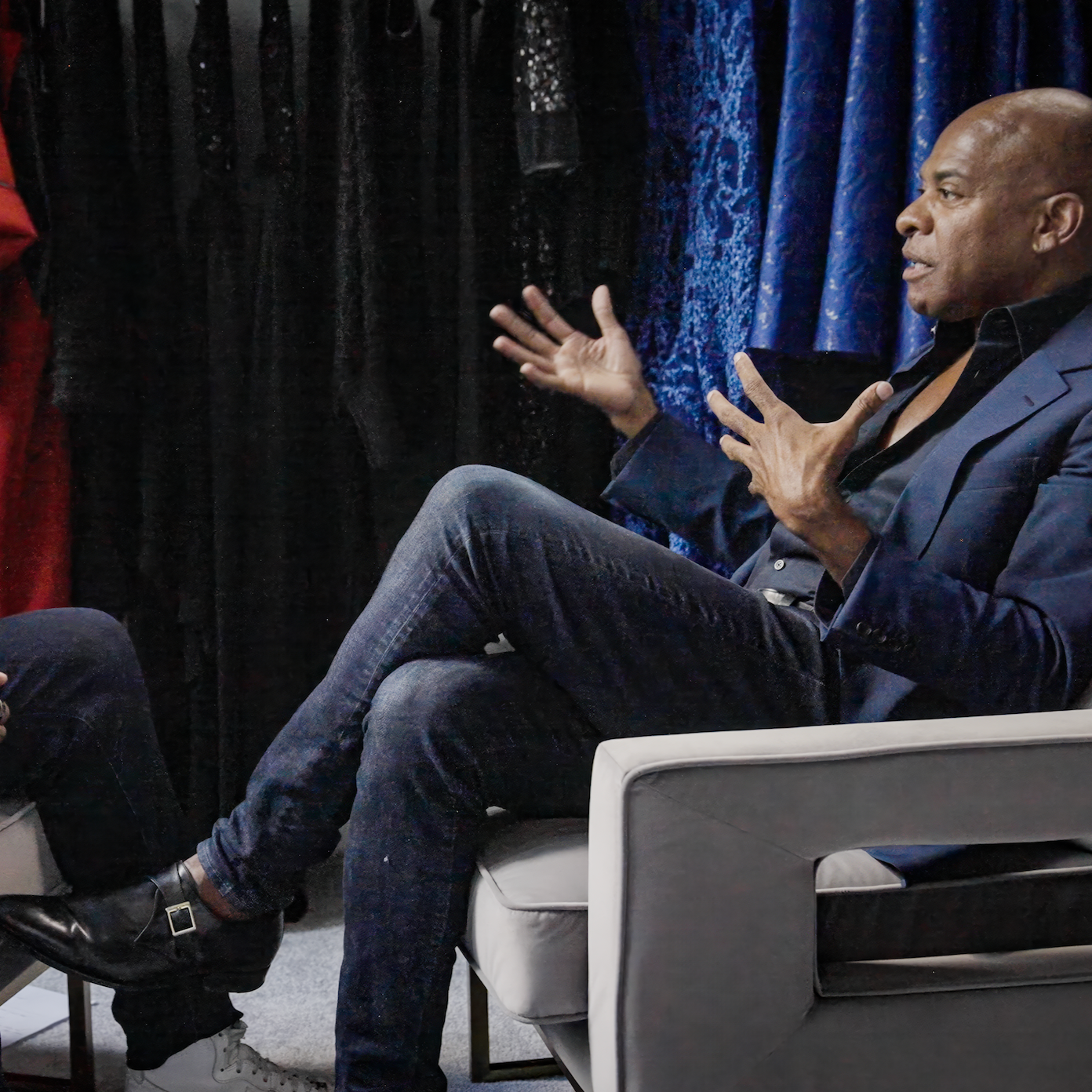 A man in a dark suit and open collar shirt sitting on a light-colored chair, gesturing with his hands while speaking, with a blue curtain and black background behind him.