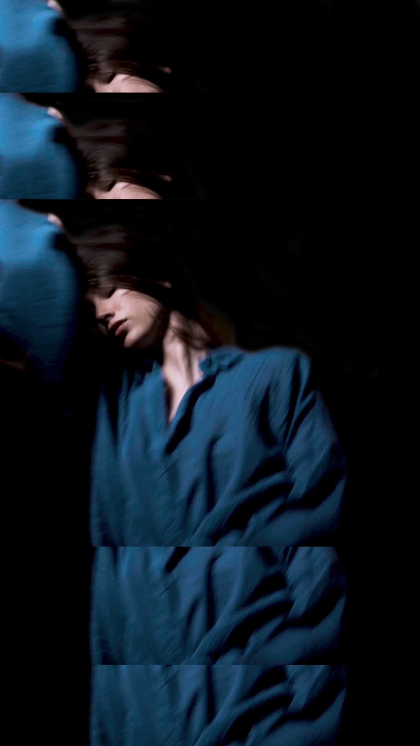 A woman with long brown hair wearing a loose blue shirt, lying down with her eyes closed, in a dark setting.