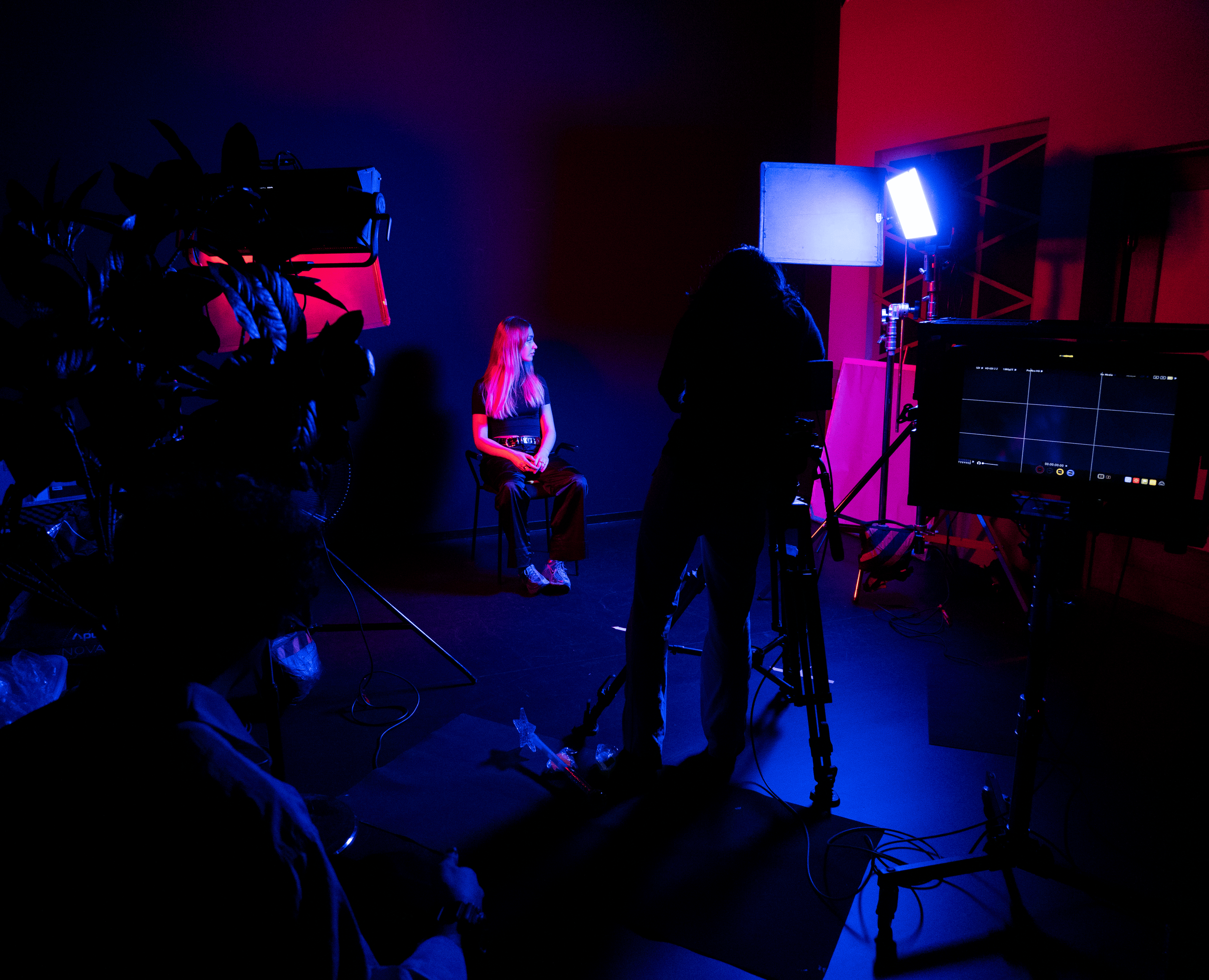 A woman with long blonde hair sitting on a chair during a photo or video shoot in a low-lit studio with colorful blue and pink lighting, a photographer standing nearby, and a monitor displaying the shoot.