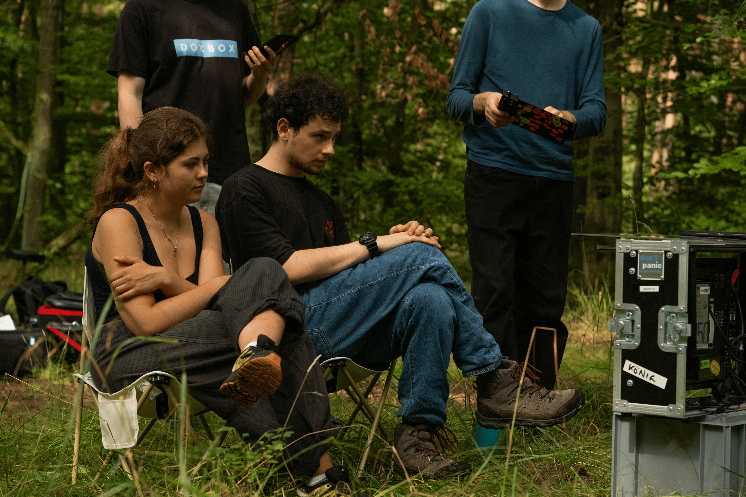 Four young adults in a forest setting, two seated on camping chairs and two standing, with filming equipment around them.