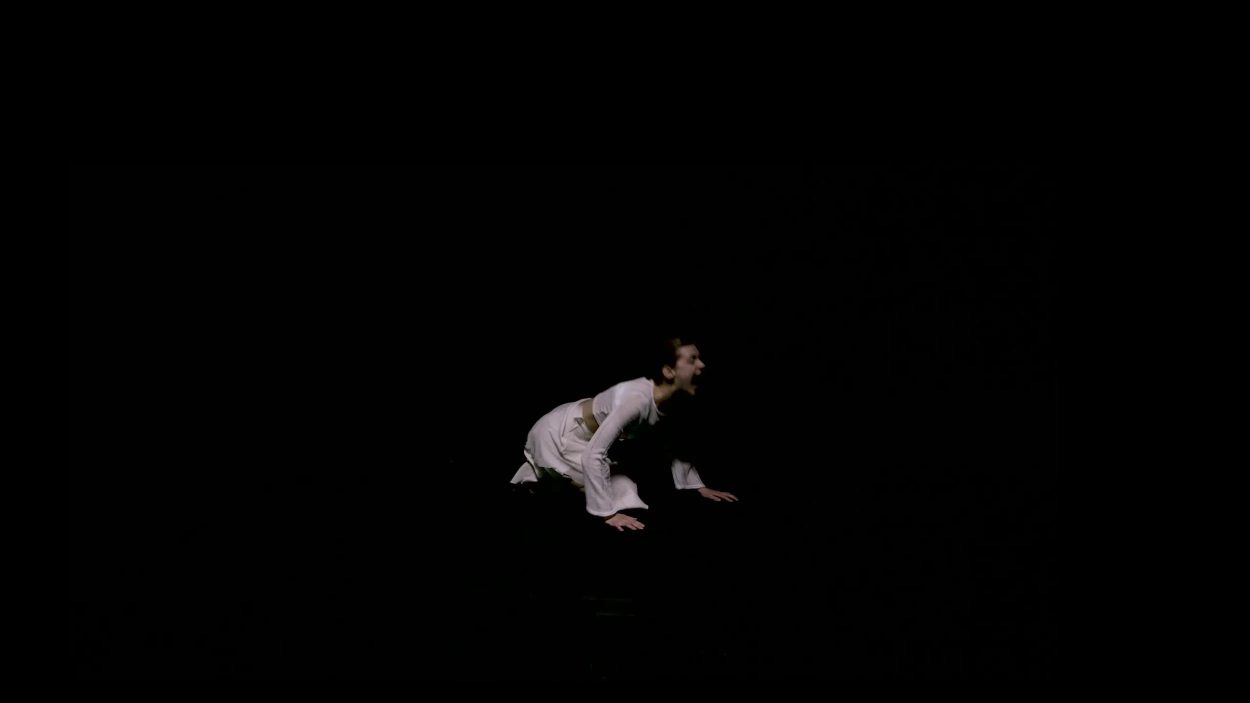 A woman in a white martial arts uniform crouches on the floor in a theatrical pose, with her mouth open, against a completely black background.
