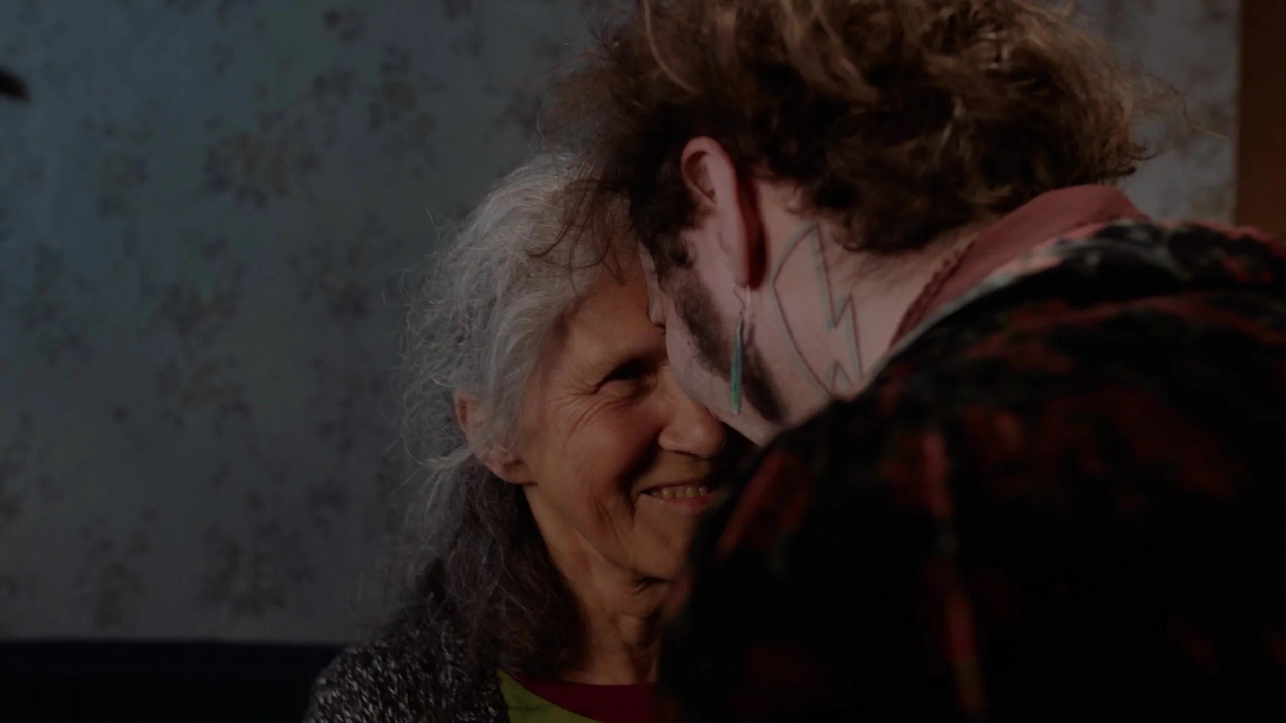A woman with gray hair and a man with a painted face are smiling and touching foreheads closely.