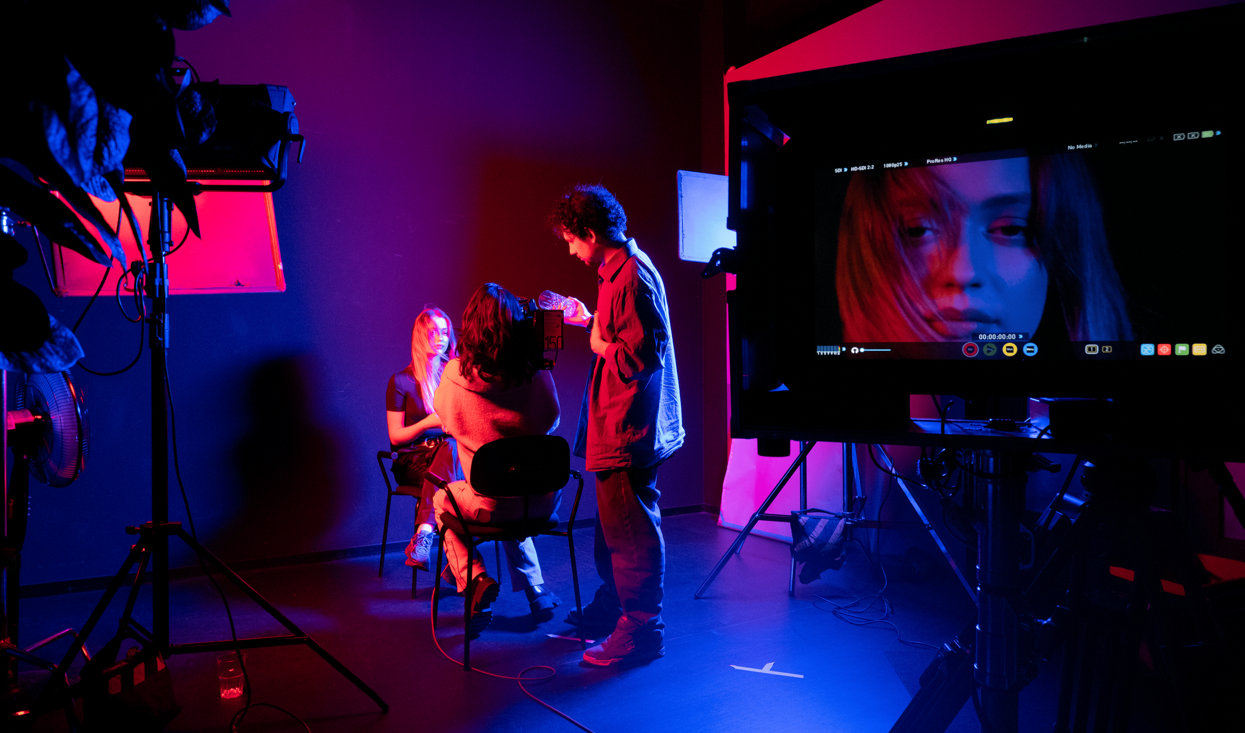 A behind-the-scenes shot of a photo or video shoot in a studio with purple and pink lighting. Two women are seated, and one man appears to be adjusting something for the shoot. There is a large monitor displaying a close-up of a woman's face.
