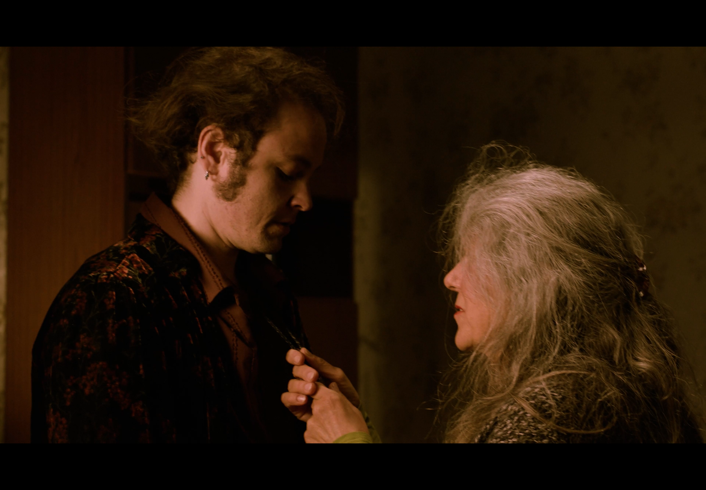 A young man with brown, curly hair and a beard wearing a dark, patterned shirt looking down at an elderly woman with long gray hair, who is holding a necklace close to his chest in a dimly lit room.