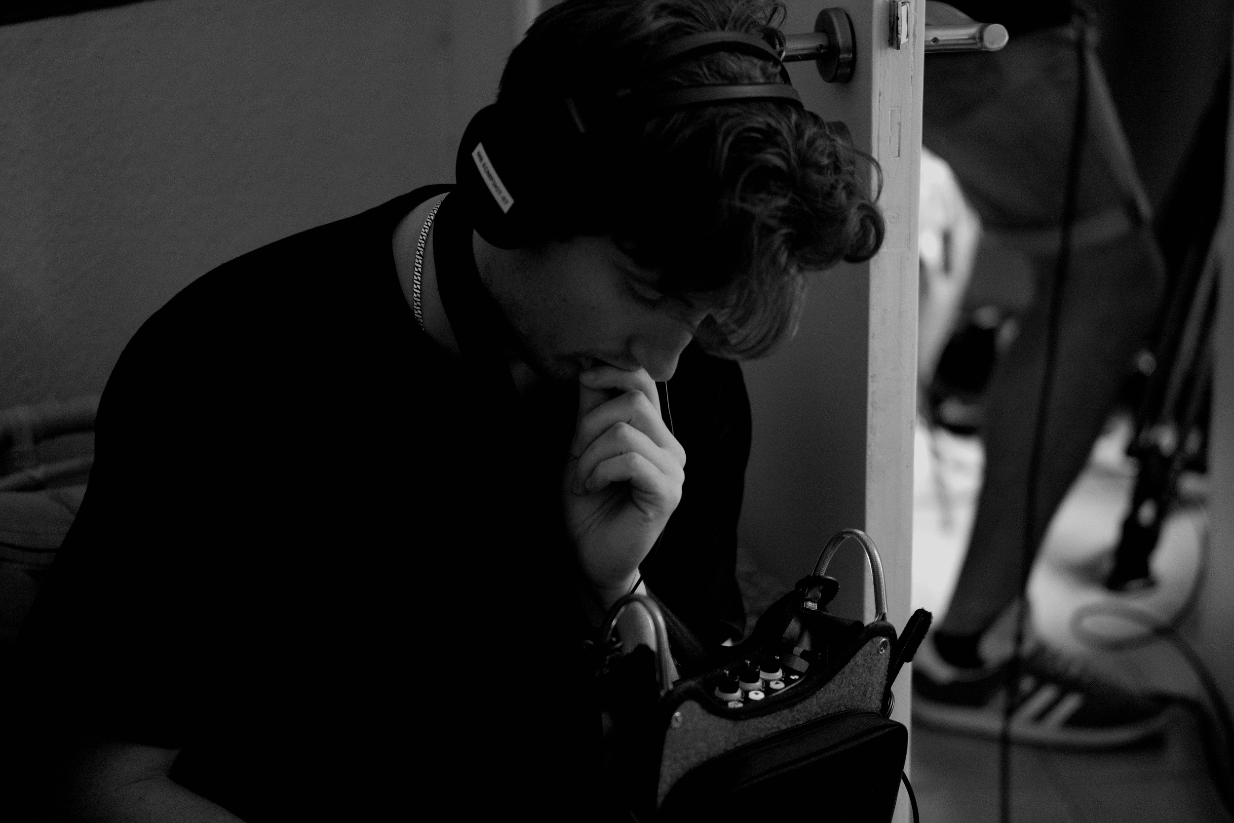 A man wearing headphones, looking down thoughtfully, sitting beside a small audio equipment case.