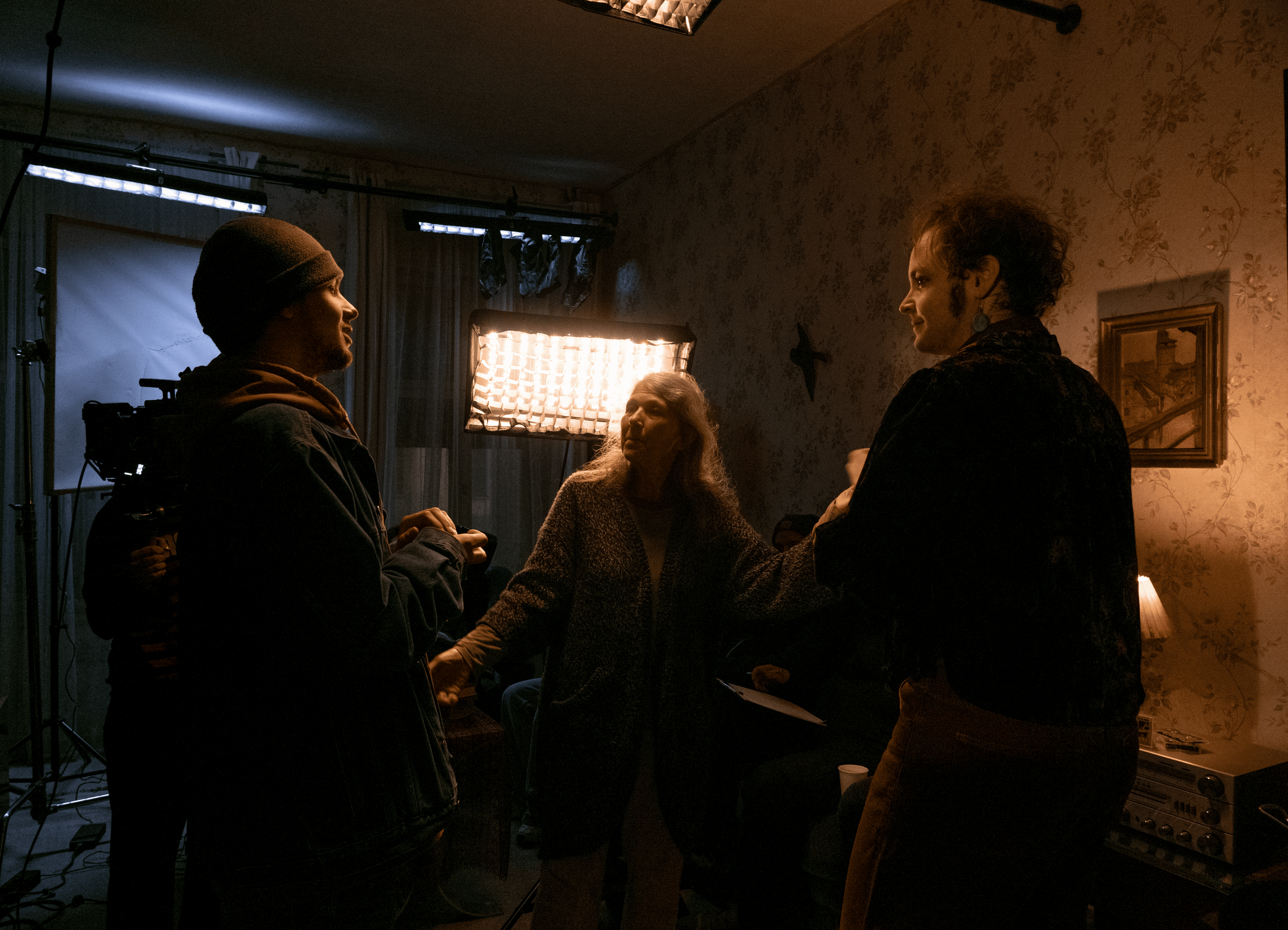 Filmmaking crew members on set, illuminated by studio lights, engaged in conversation in a dimly lit room with patterned wallpaper, framed picture, and film equipment.
