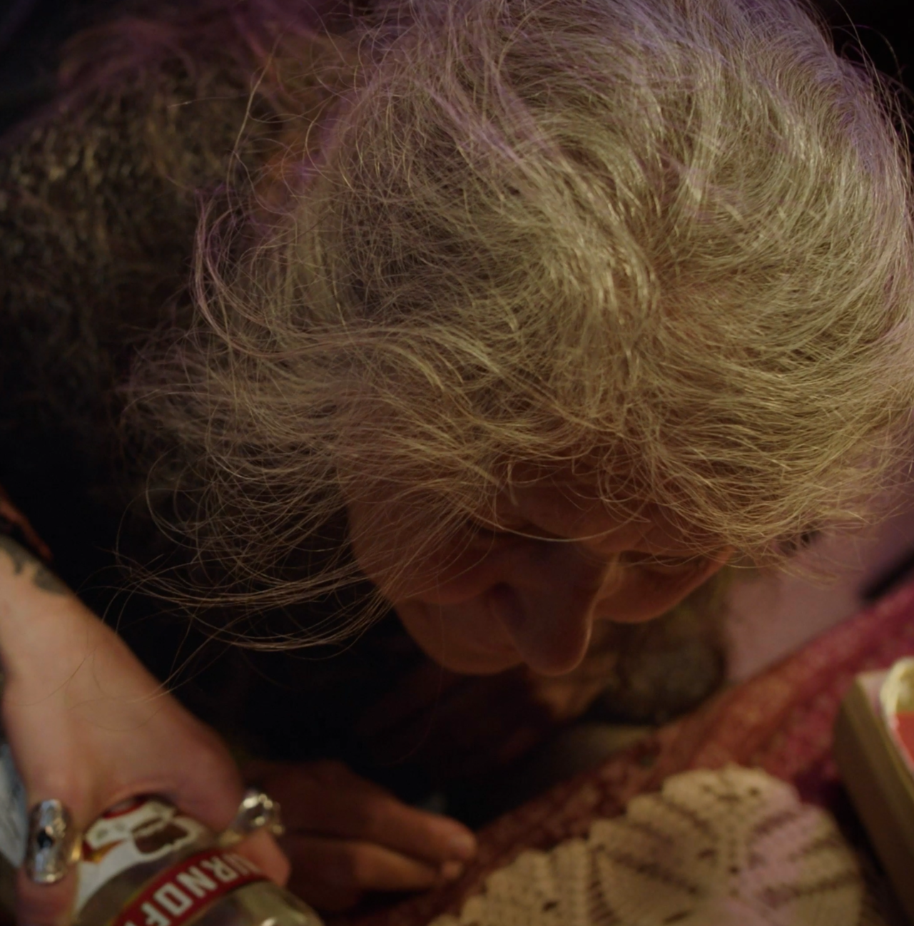 Close-up of an elderly woman's face with curly grey hair, looking downward, with her hand holding a glass of alcohol, next to a plate of food on a table.