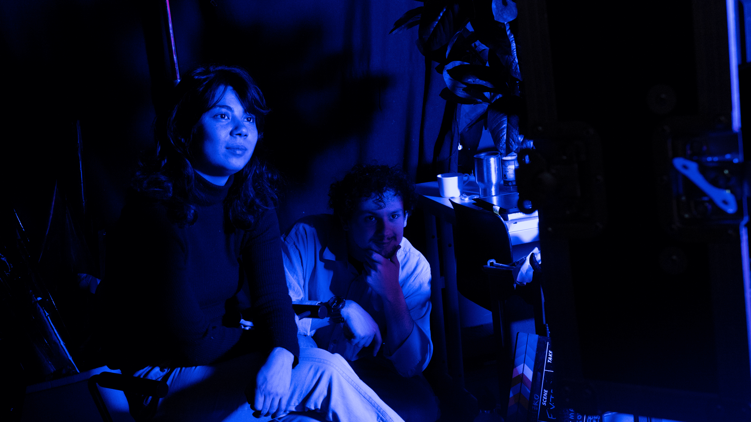 Two people, a woman with dark wavy hair and a man with curly hair, sitting together in a room lit with blue light, surrounded by film equipment and a clapperboard on the floor.