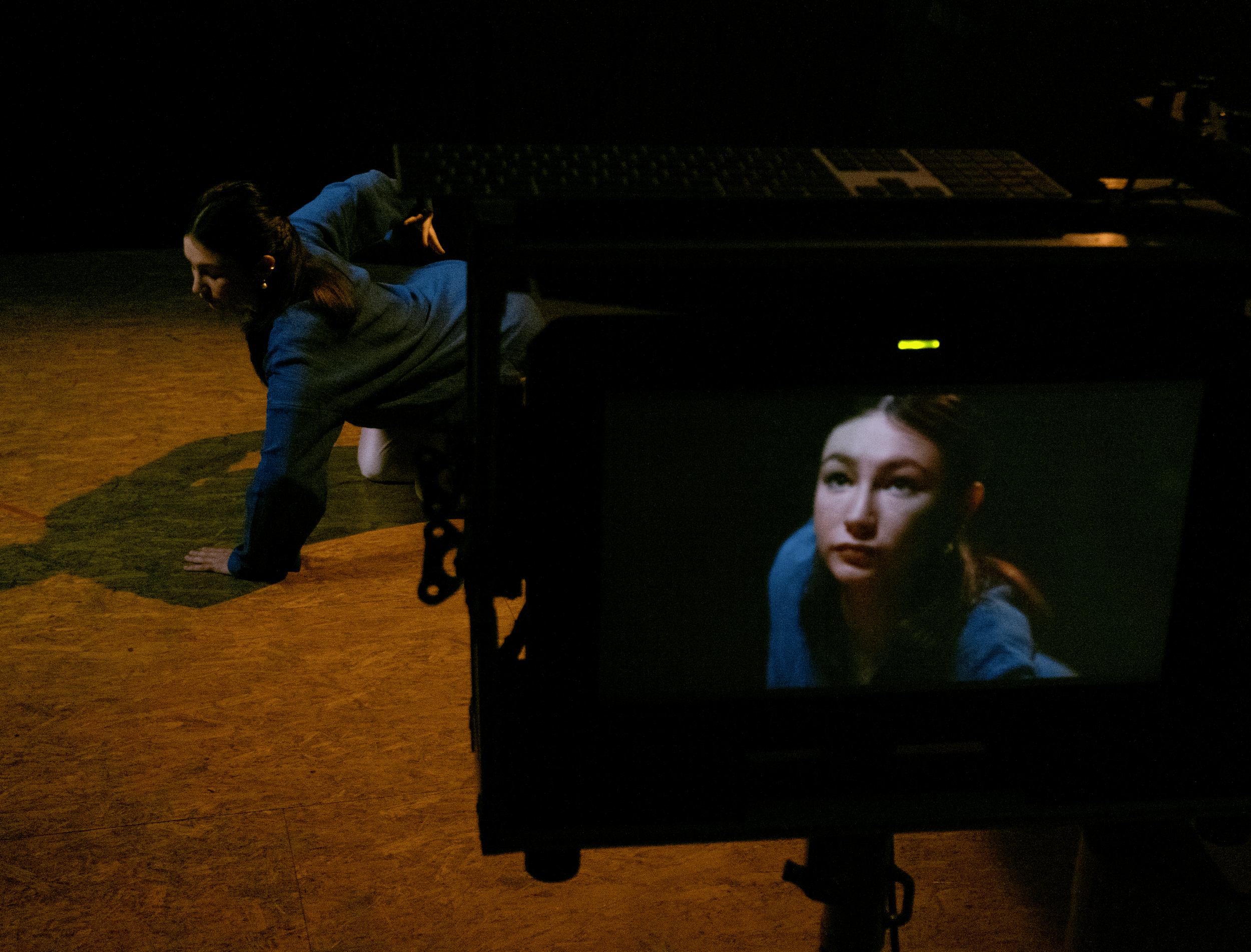 A woman in a blue shirt is performing a yoga pose on a wooden floor, with her one hand on the ground and her body extended sideways. There is a camera filming her, and the camera's monitor shows a close-up of her face.