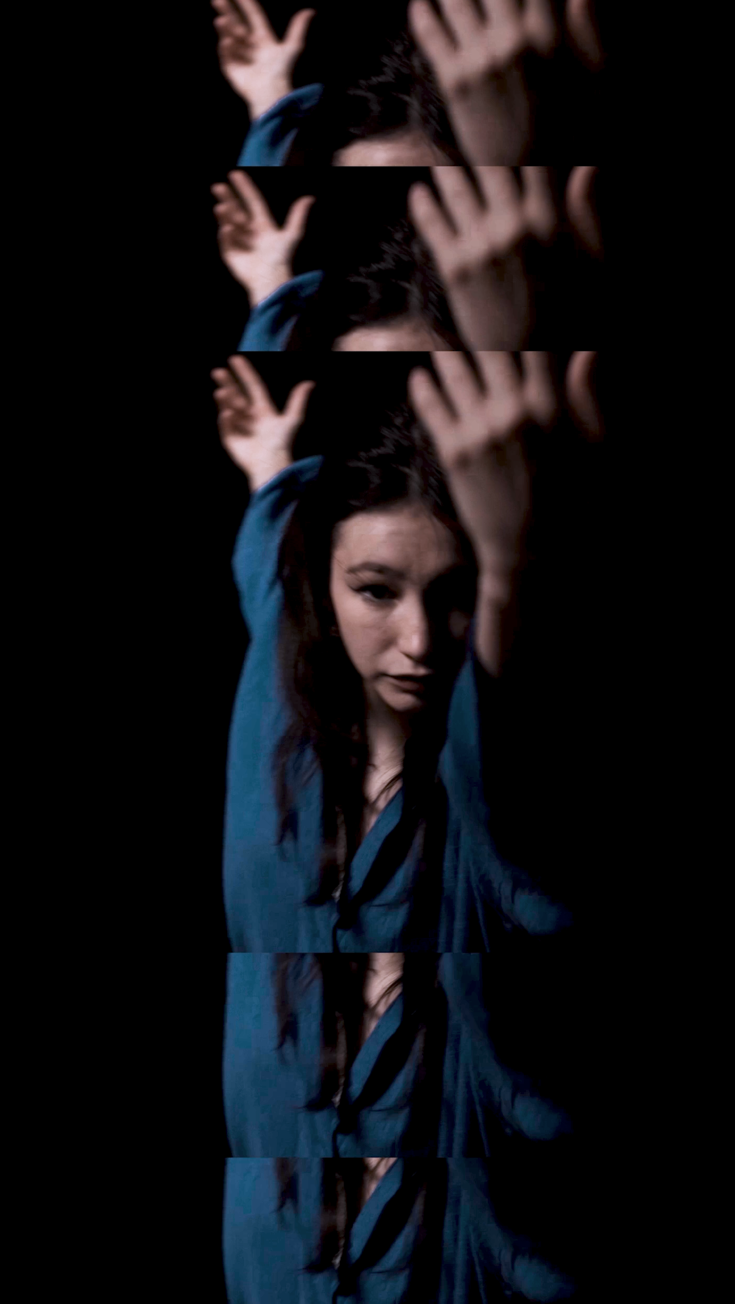 A young woman with long dark hair and a blue shirt, raising her hands above her head in a pose against a dark background.