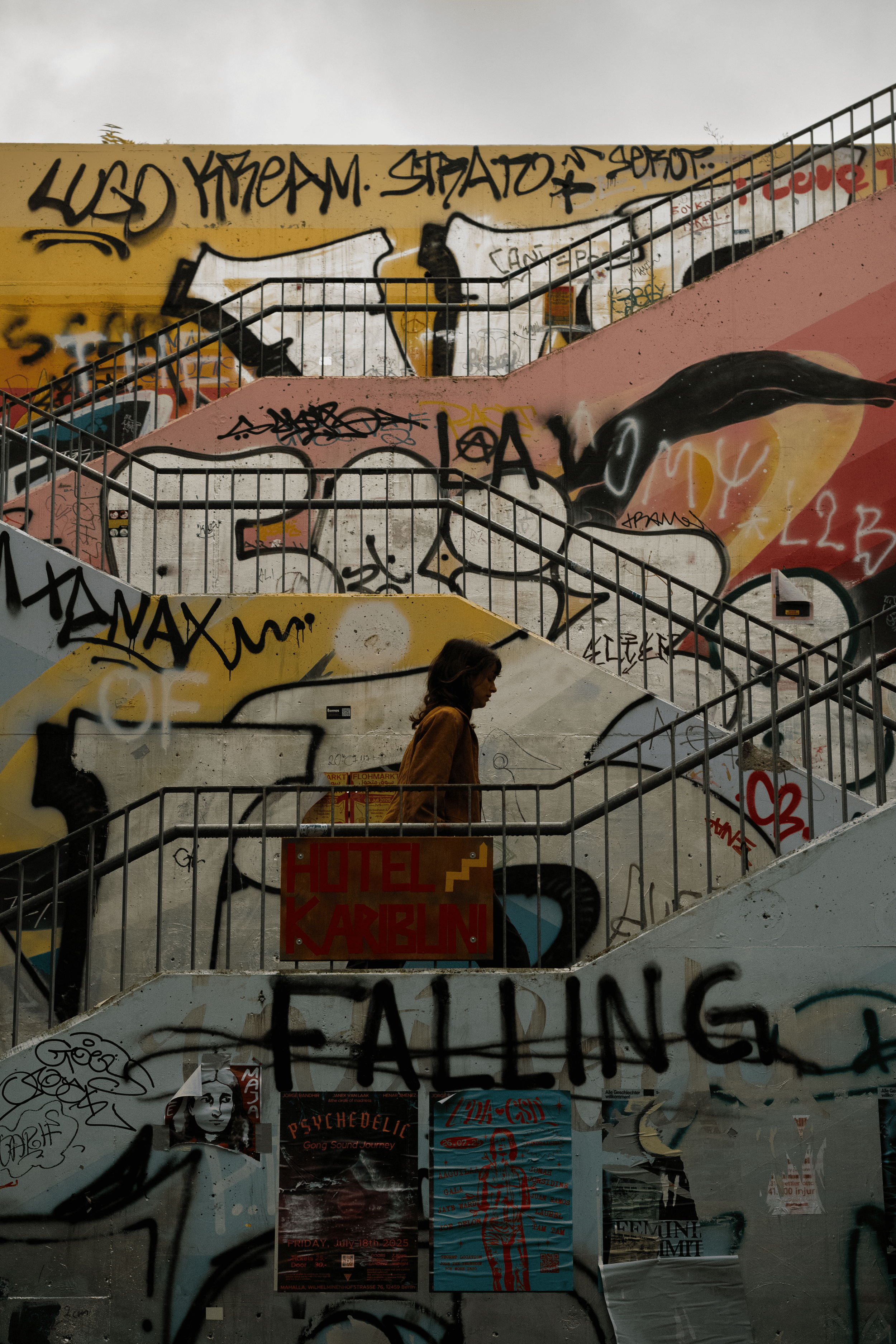 A woman walking up a graffiti-covered outdoor staircase with multiple landings and railings, and signs with graffiti art and posters on the walls.