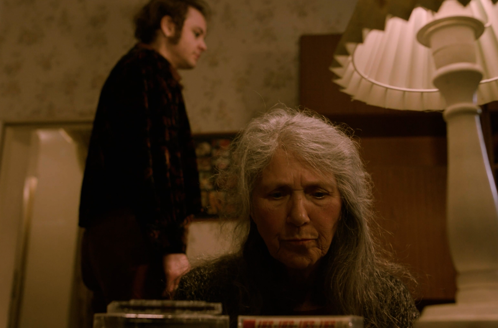 A woman with long gray hair sitting at a piano, looking down with a serious expression, while a man with curly hair and sideburns stands in the background, looking away in a dimly lit room.