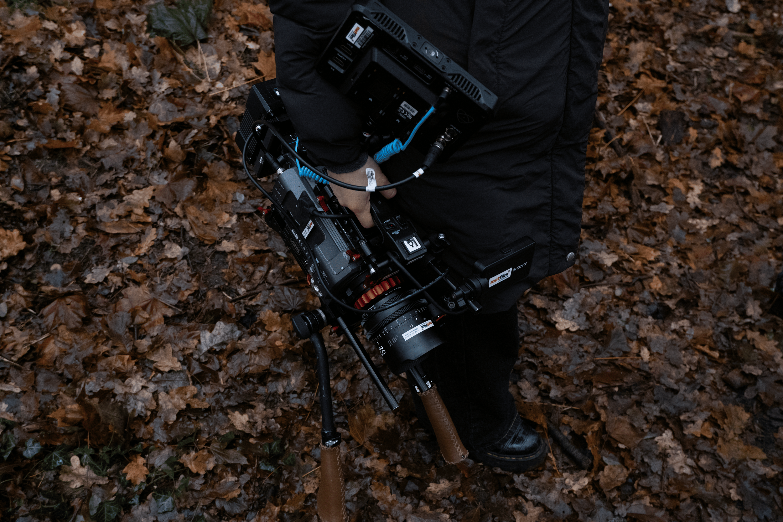 Close-up of a person holding a professional film or video camera with various attachments, standing on fallen autumn leaves.