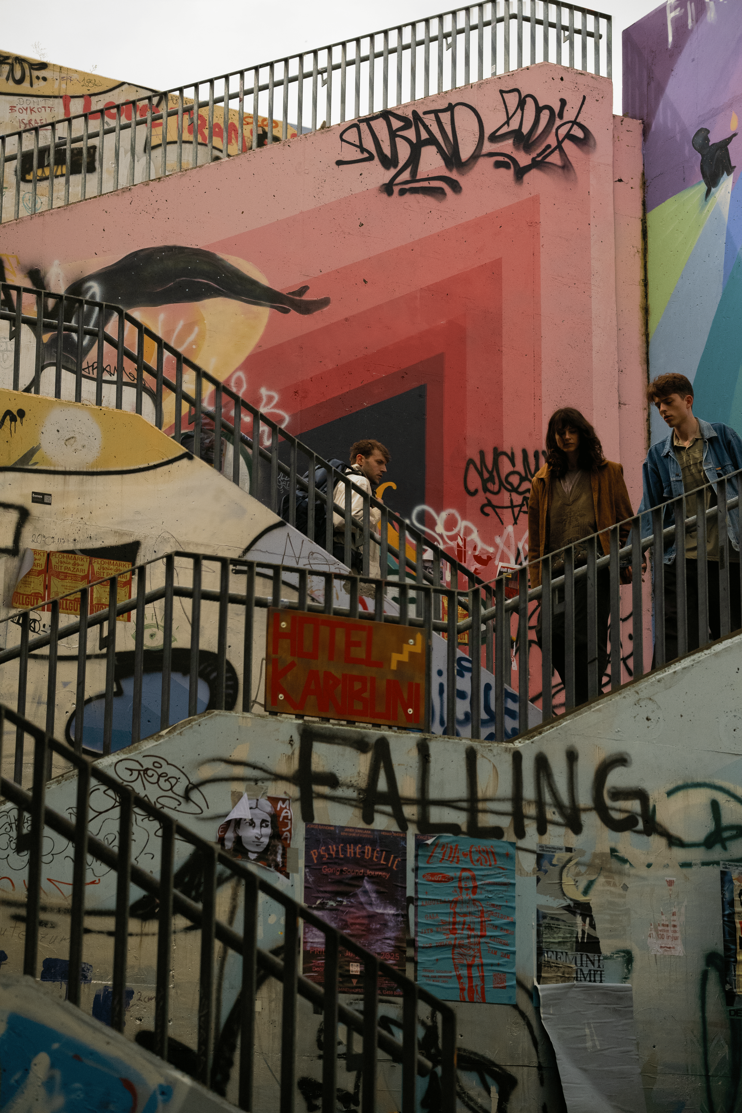 People walking down an outdoor stairway covered in graffiti and posters, with a colorful mural in the background.