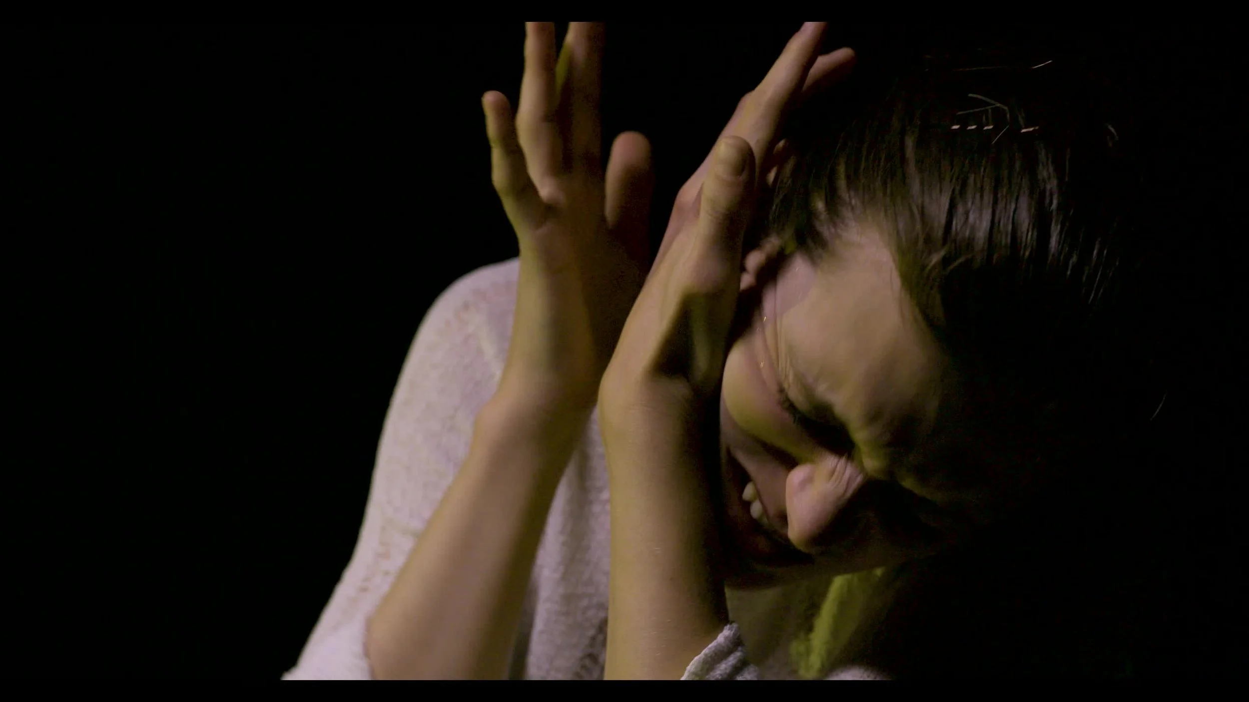 A woman appears distressed, covering her ears with her hands and grimacing, with a dark background.