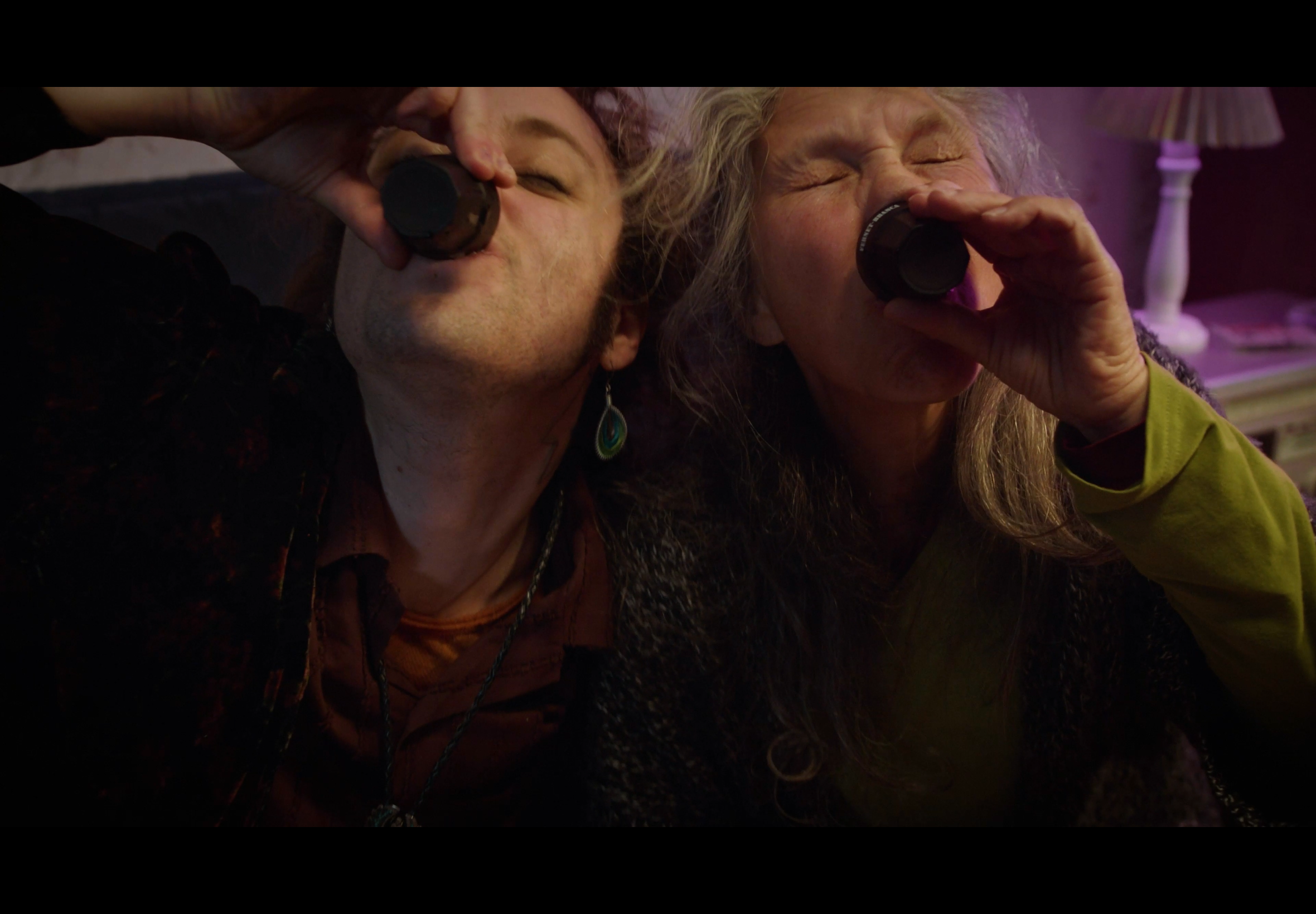 Two older women drinking from small bottles, sitting closely together indoors with a lamp and a pinkish background.
