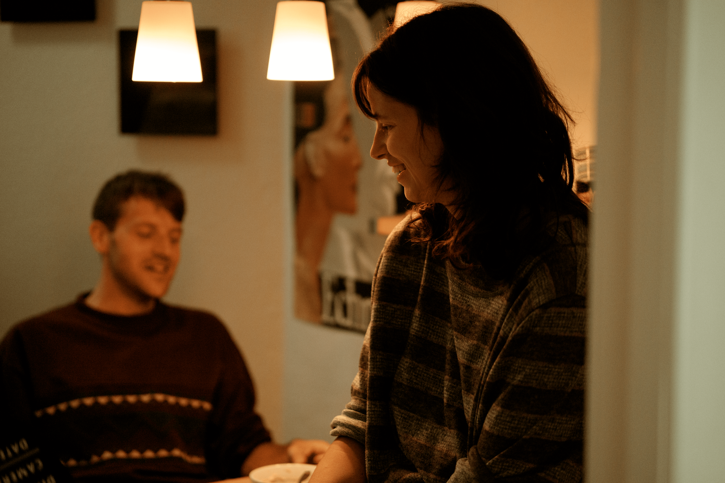 A woman with dark hair smiling and looking down, with a young man with short hair in the background, both indoors under warm lighting.