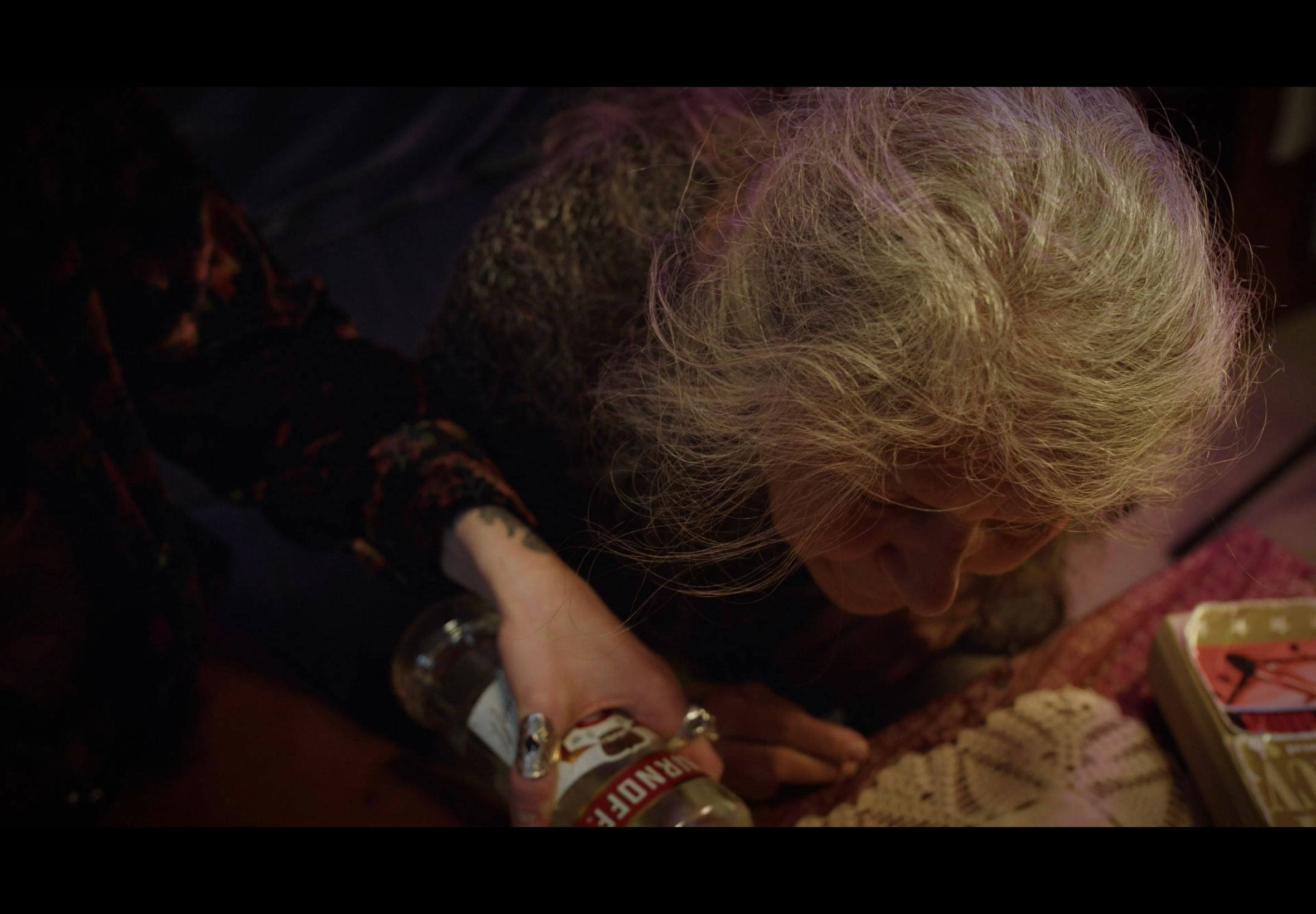 A woman with curly blonde hair sitting on the floor, leaning over a table with items including a bottle of Jack Daniel's whiskey and a box of matches.