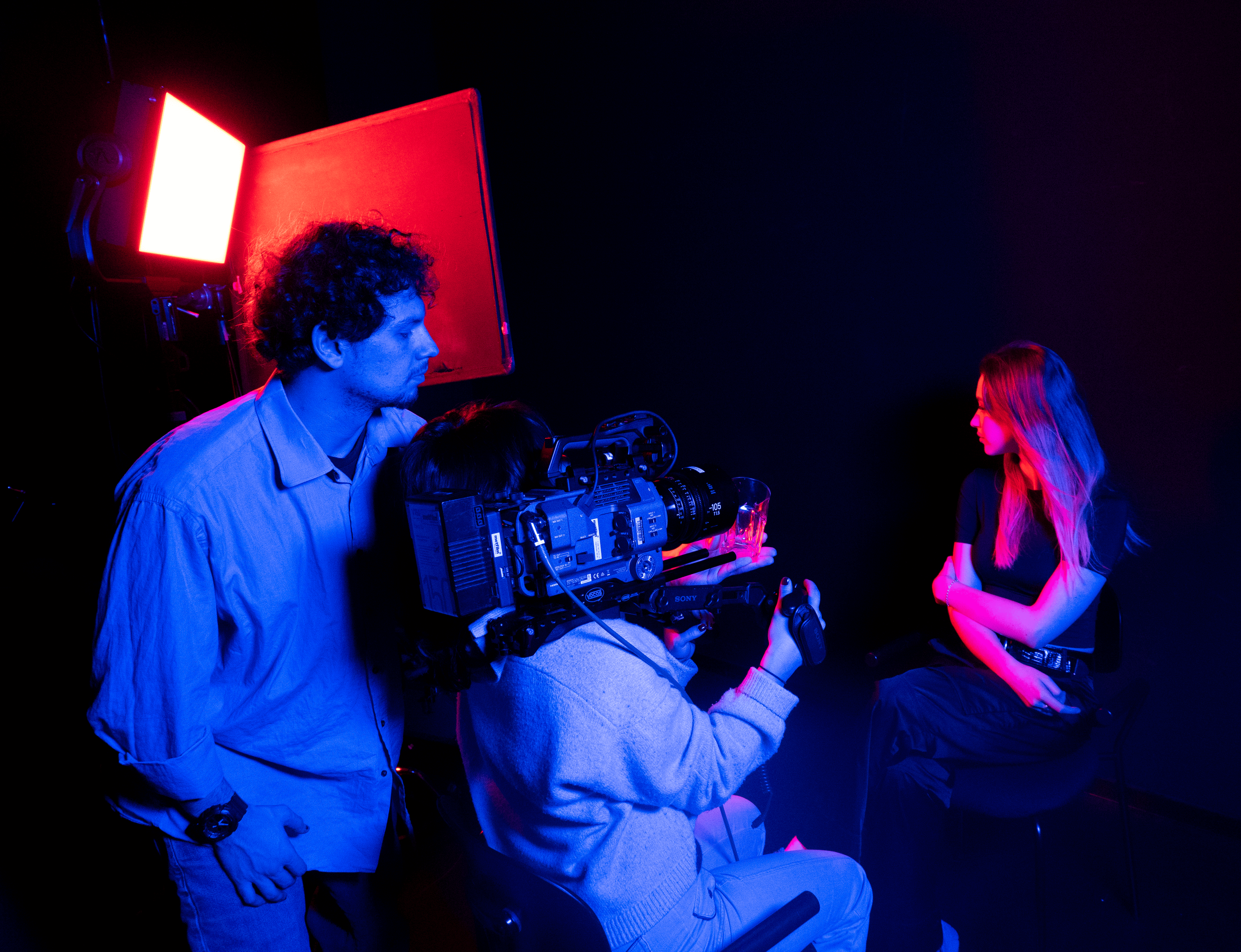 A behind-the-scenes view of a video shoot with a woman sitting on a chair, surrounded by two crew members in a dark studio illuminated by red and blue lighting. One crew member is operating a professional camera, while the other stands nearby.