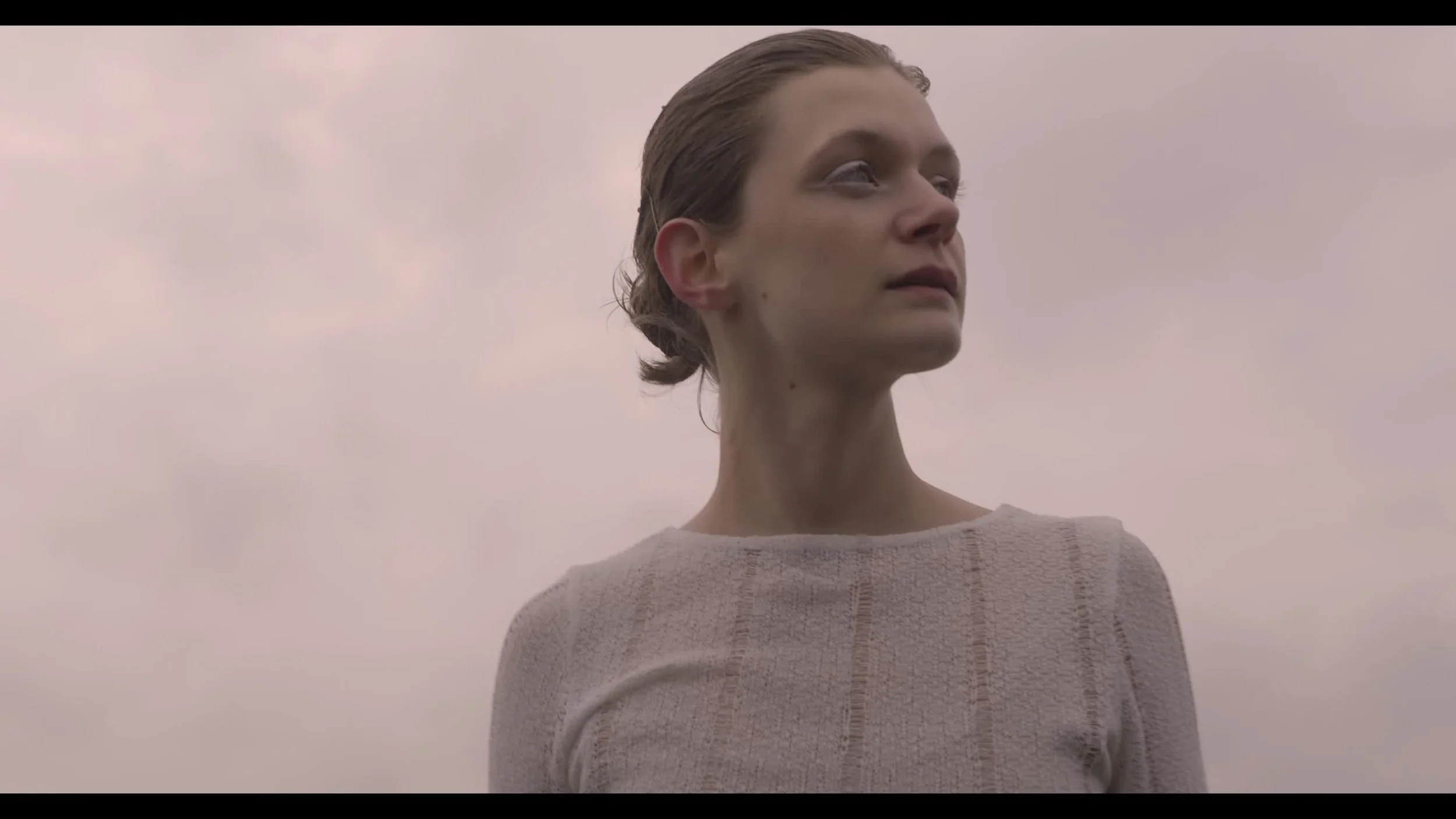A woman with her hair tied back, looking into the distance against a cloudy sky.