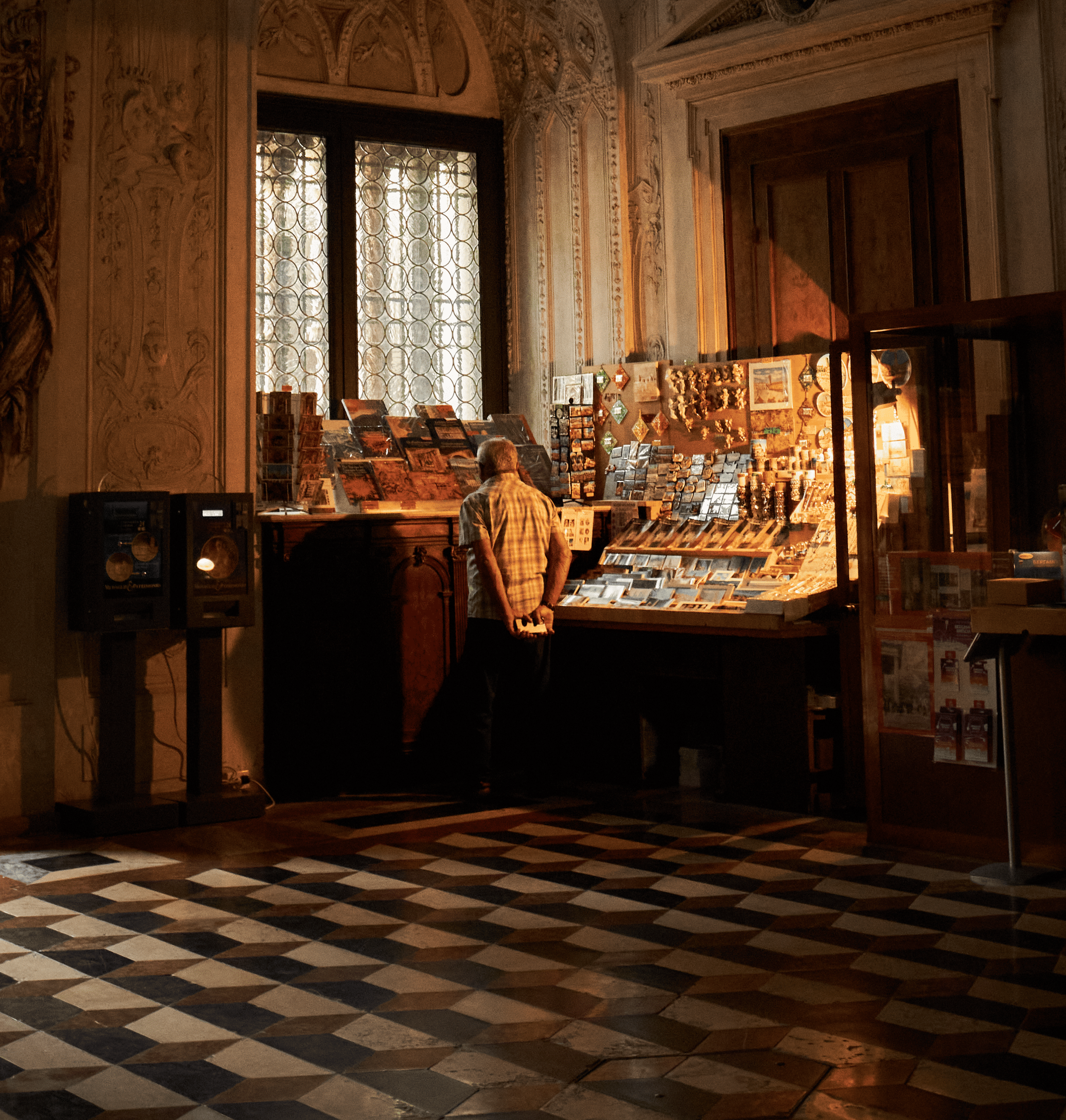 An elderly man in a checked shirt browsing postcards and souvenirs at a display stand inside a historic building with ornate walls and patterned tiled floors.