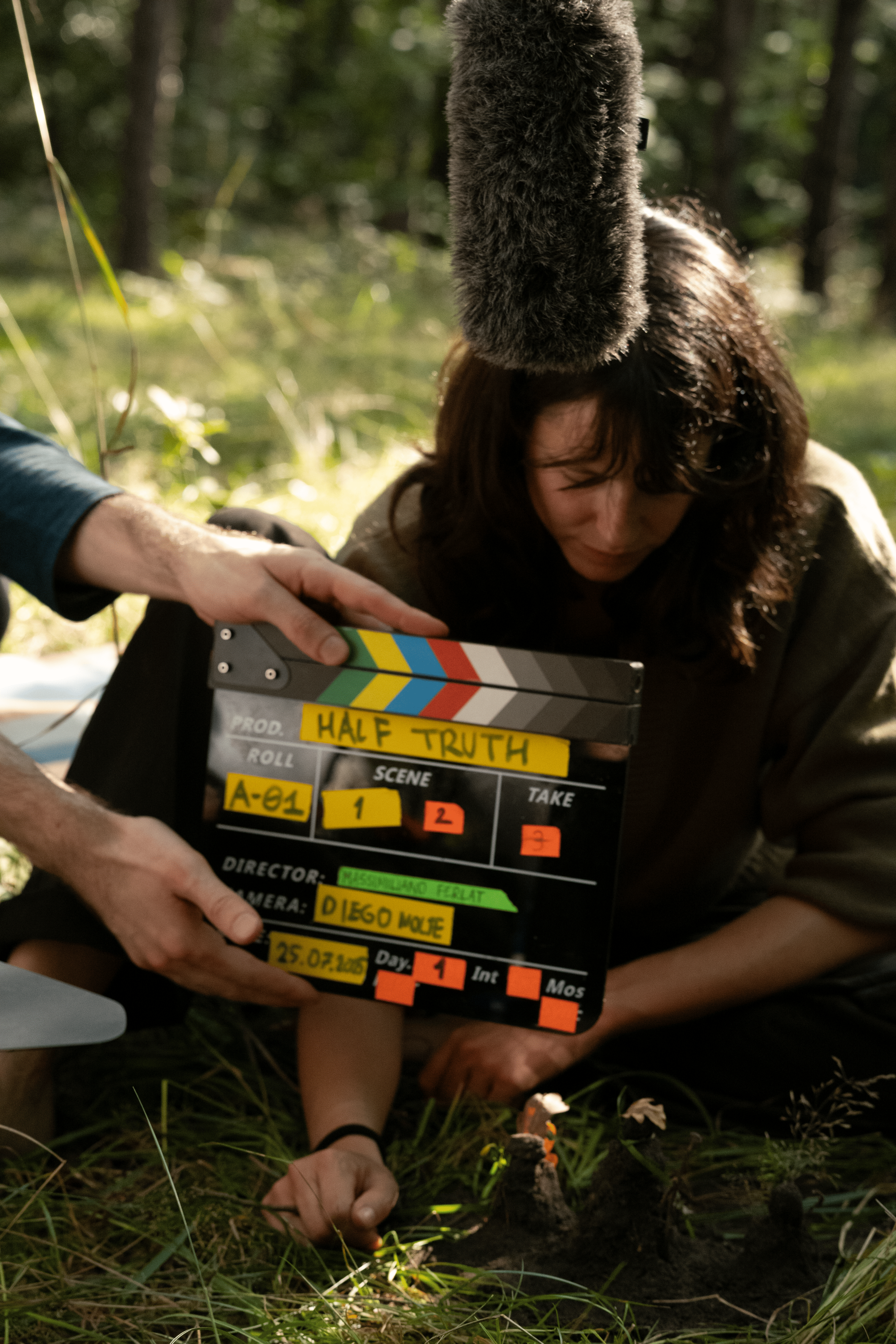 A woman is lying on the ground in a forest, with a clapperboard marked 'Half Truth' held close to her face. There is a microphone above her head, and her hand is on the grass near some small objects or plants.
