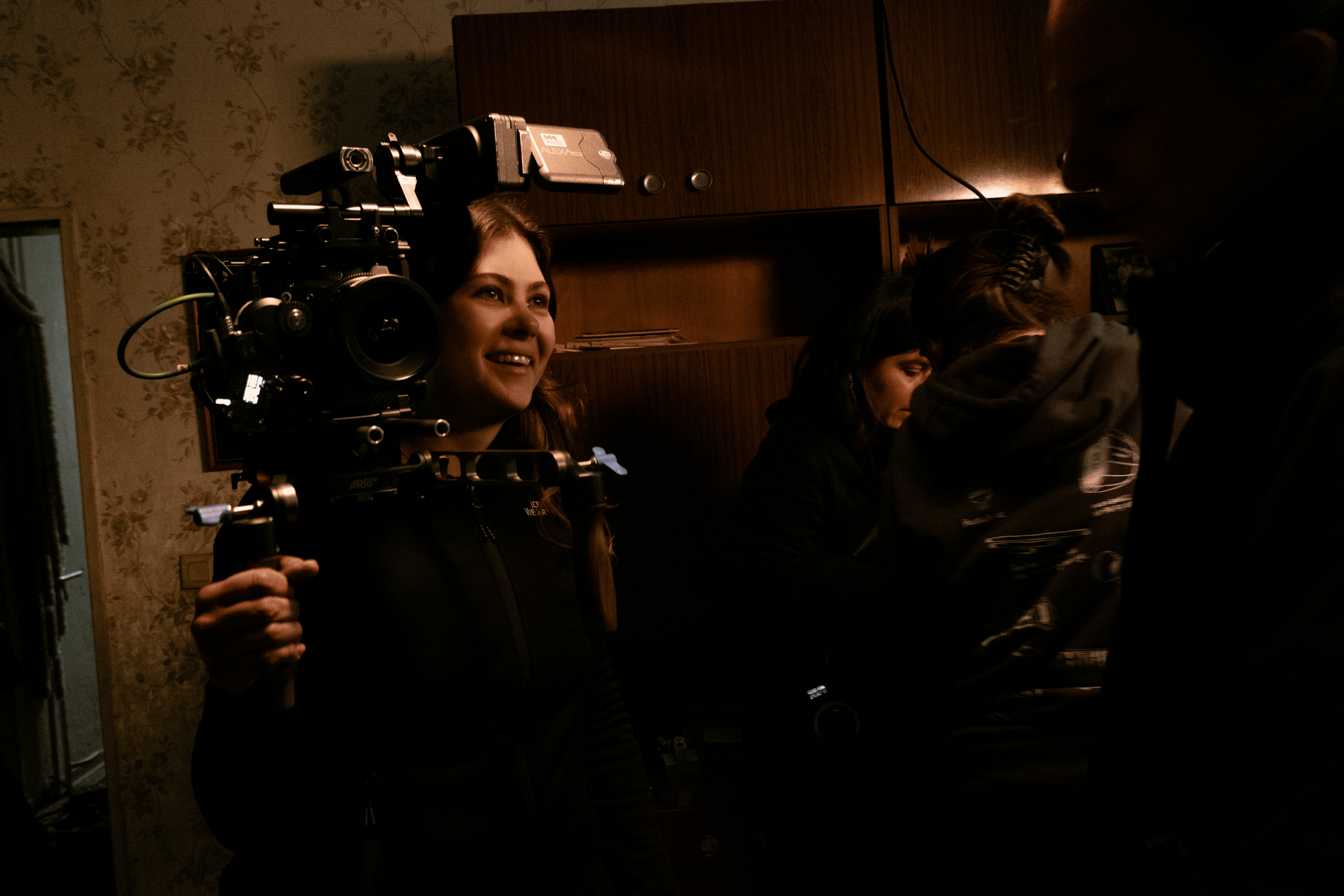 A woman holding a video camera is smiling, with several other people in the background engaged in an activity in a dimly lit room.