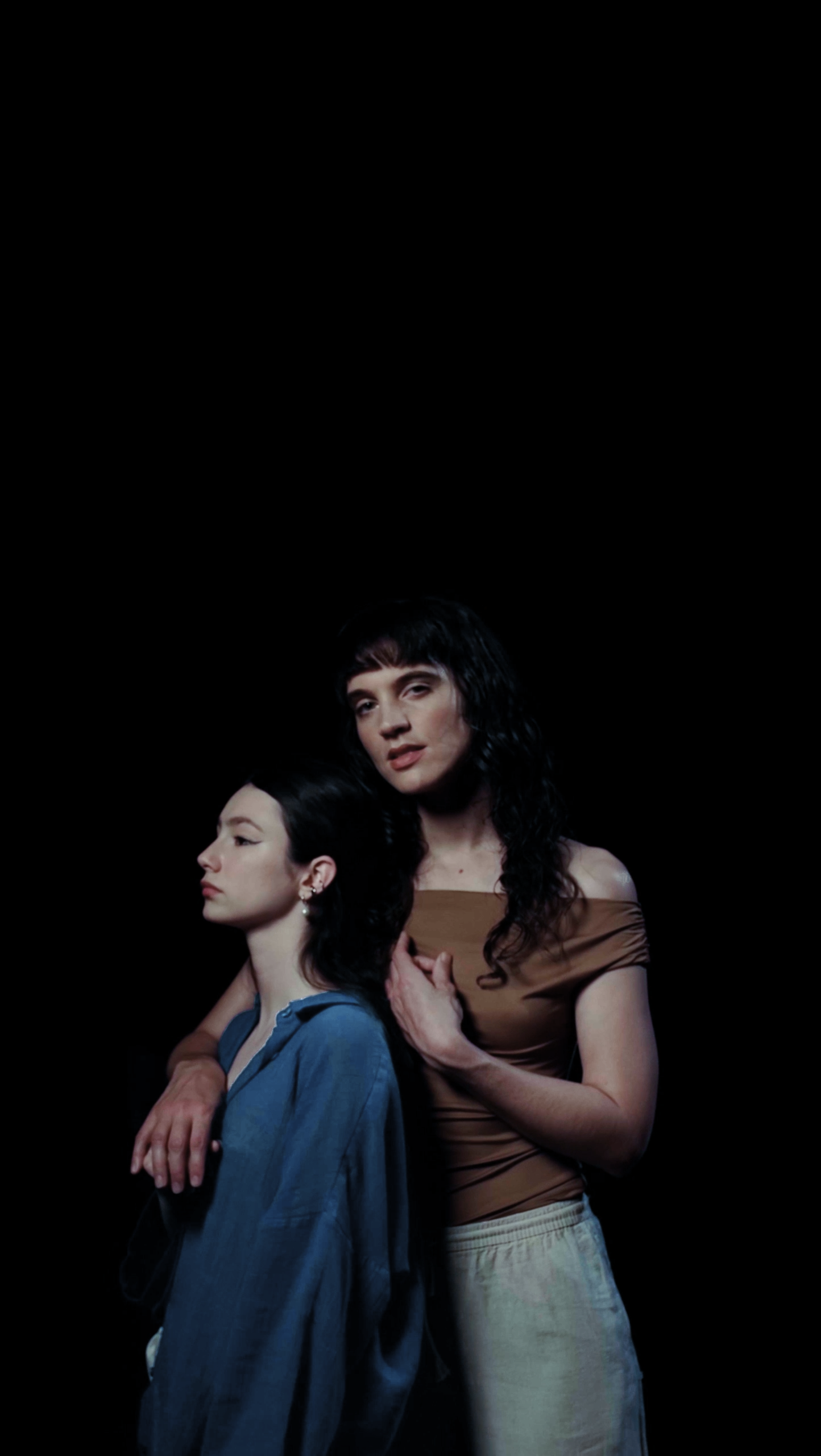 Two women pose against a black background, one with dark hair and the other with short hair and bangs. The woman with short hair looks off to the side while the other looks directly at the camera. The woman with short hair wears a blue denim shirt, a