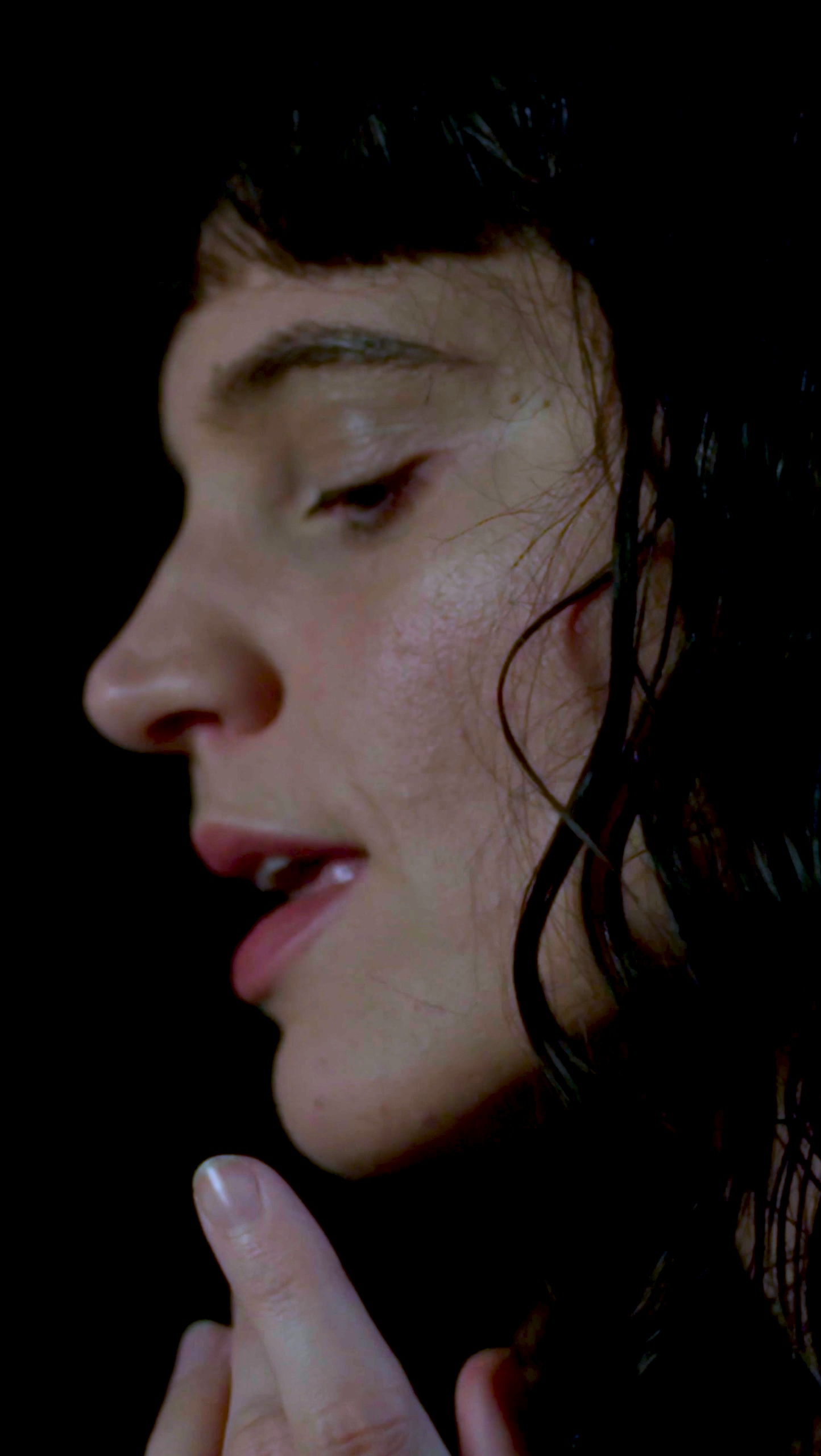 A close-up side profile of a woman with wet dark hair, light makeup, and a contemplative expression, touching her finger to her lips.